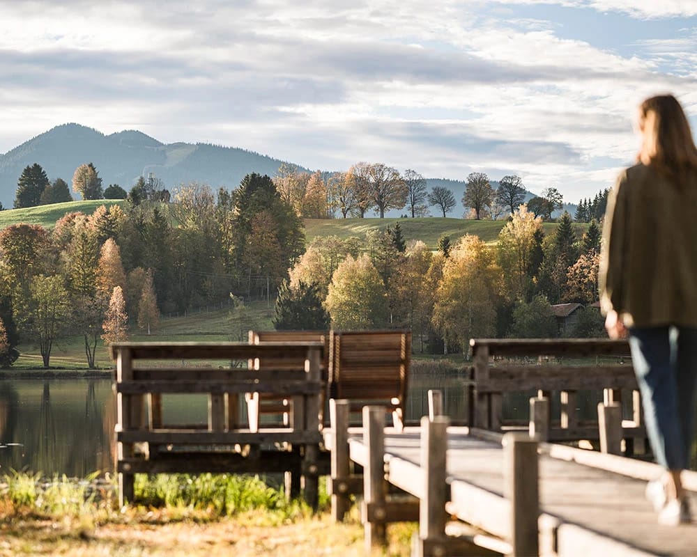 Holzsteg mit Bänken am See, dahinter herbstlich gefärbte Bäume und Hügel