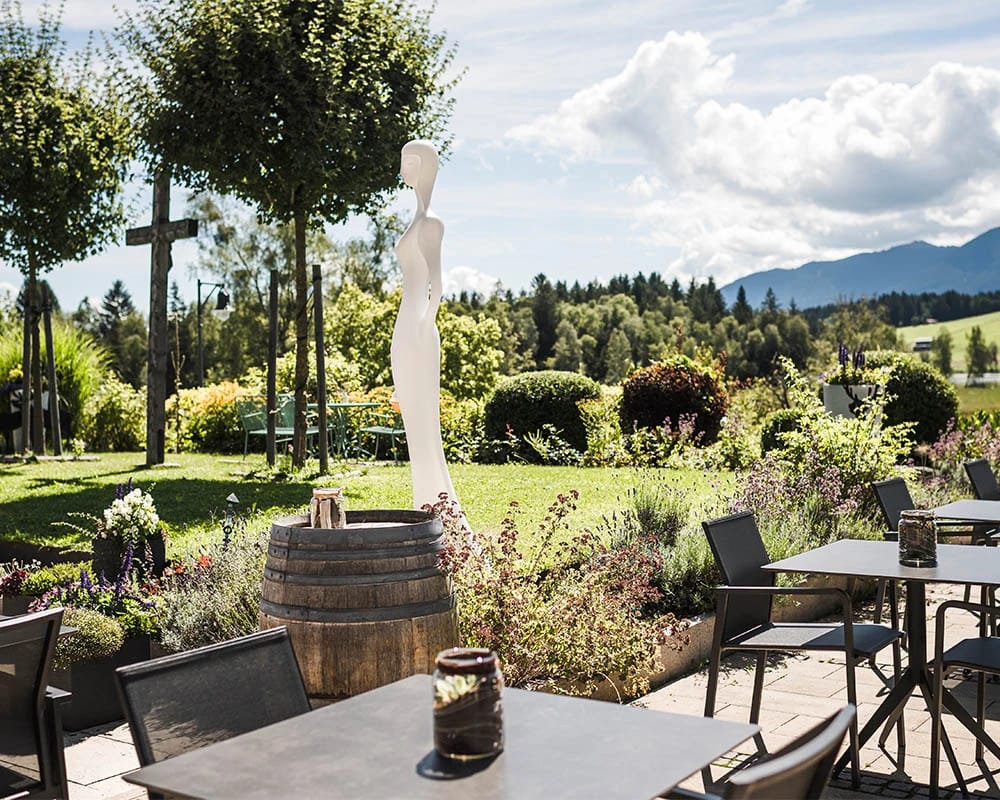 Terrasse mit Tischen, Weinfass und Skulptur im Garten, dahinter Wiesen und Berge