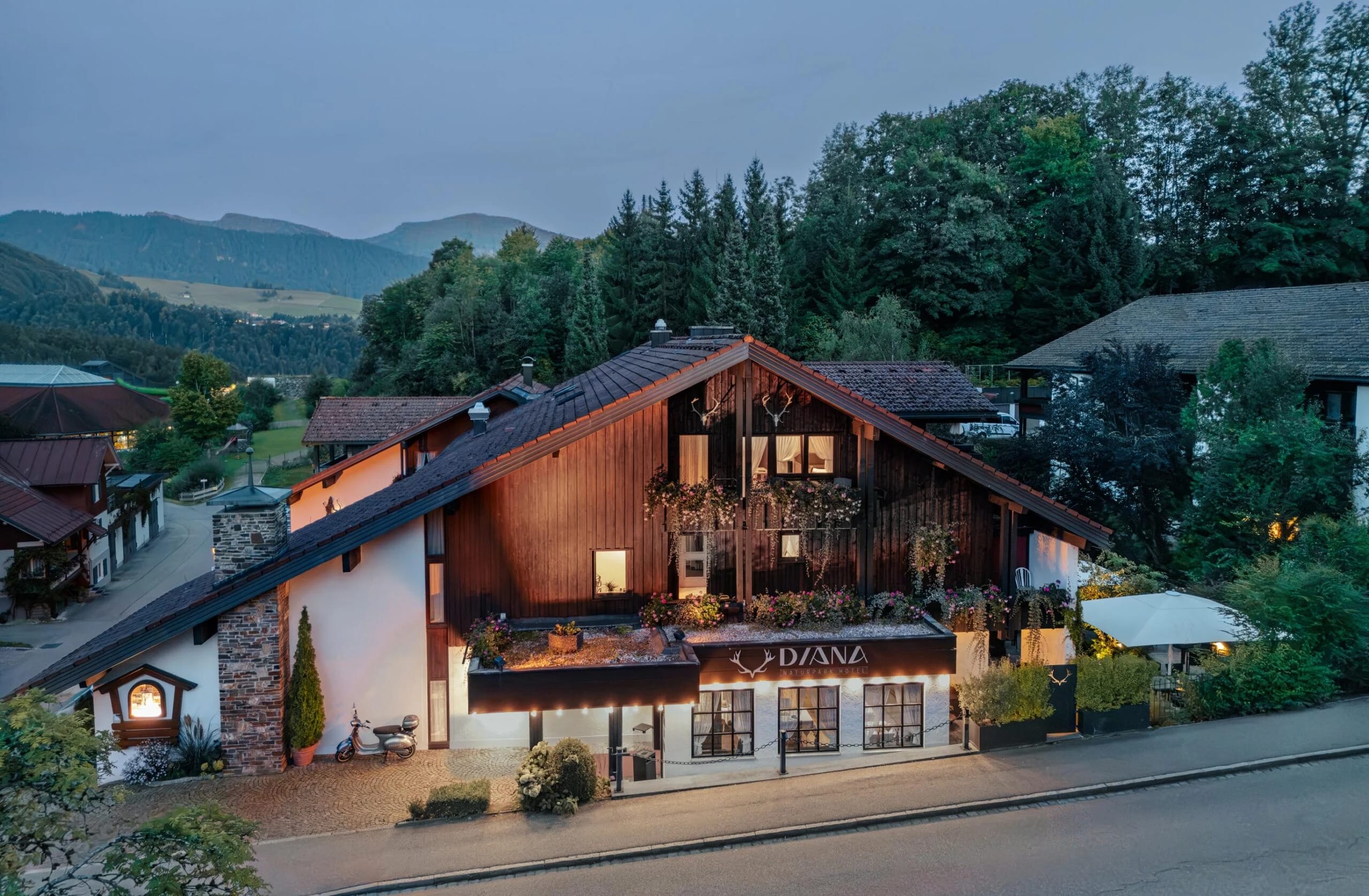 Beleuchtete Hausfassade mit Holzgiebel und Schriftzug DIANA in der Abenddämmerung, dahinter bewaldete Berge