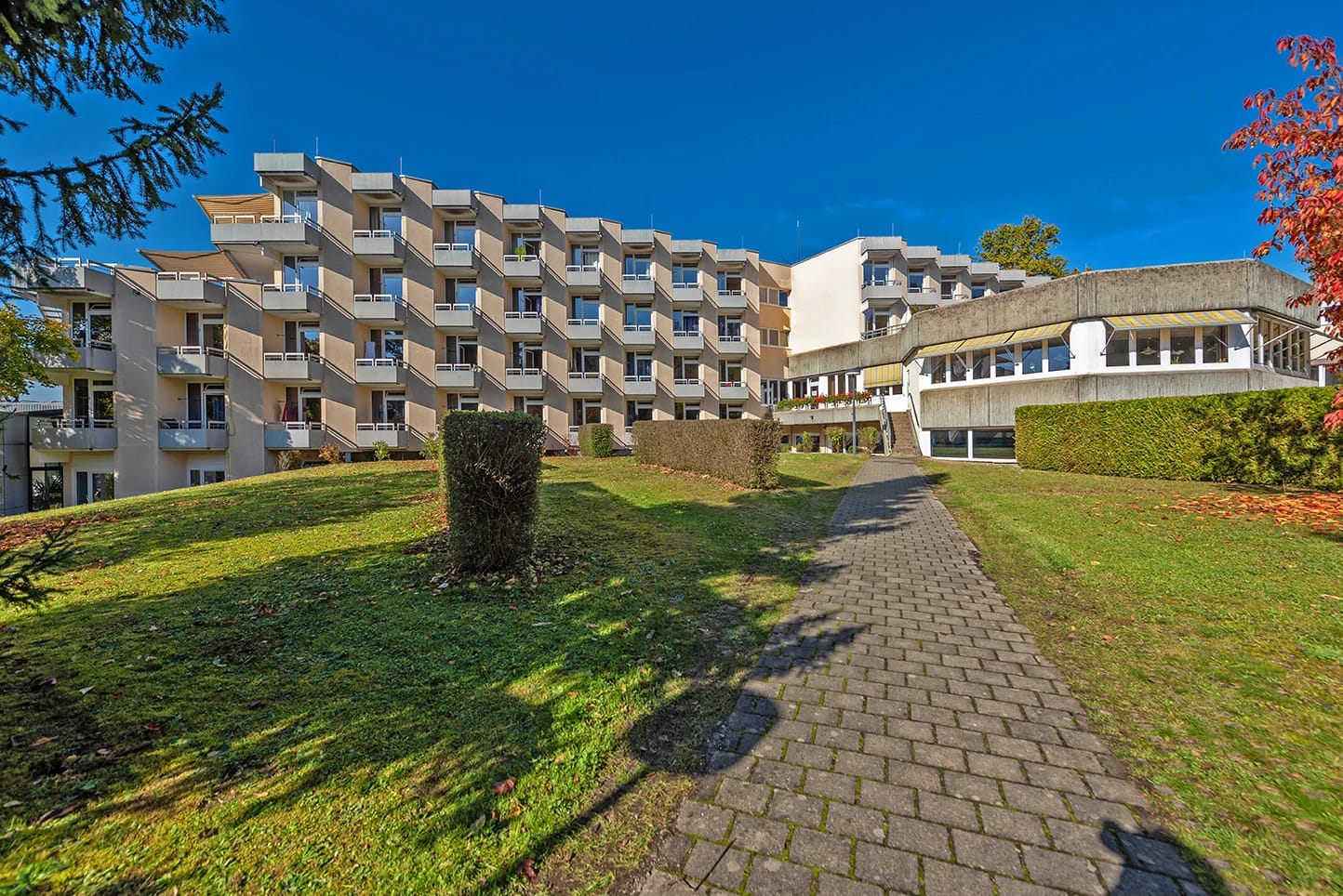Klinikgebäude mit gestaffelten Balkonen, Gehweg und Rasenfläche bei blauem Himmel