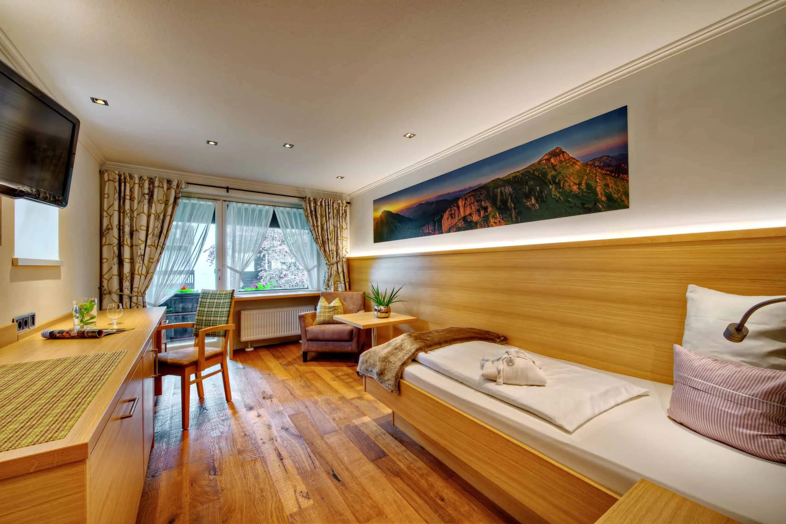 Zimmer mit Holzverkleidung, großem Bergbild über dem Bett und Sitzplatz am Fenster