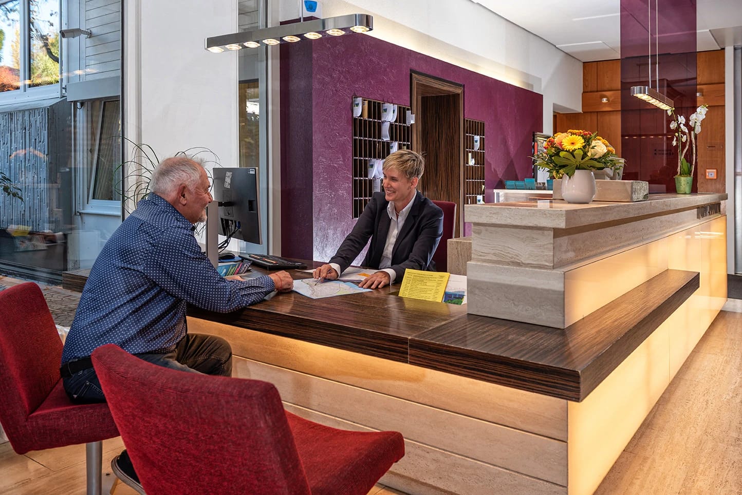 Empfangstresen aus Holz mit Mitarbeiter und Gast, Blumenschmuck und farbig abgesetzter Wand