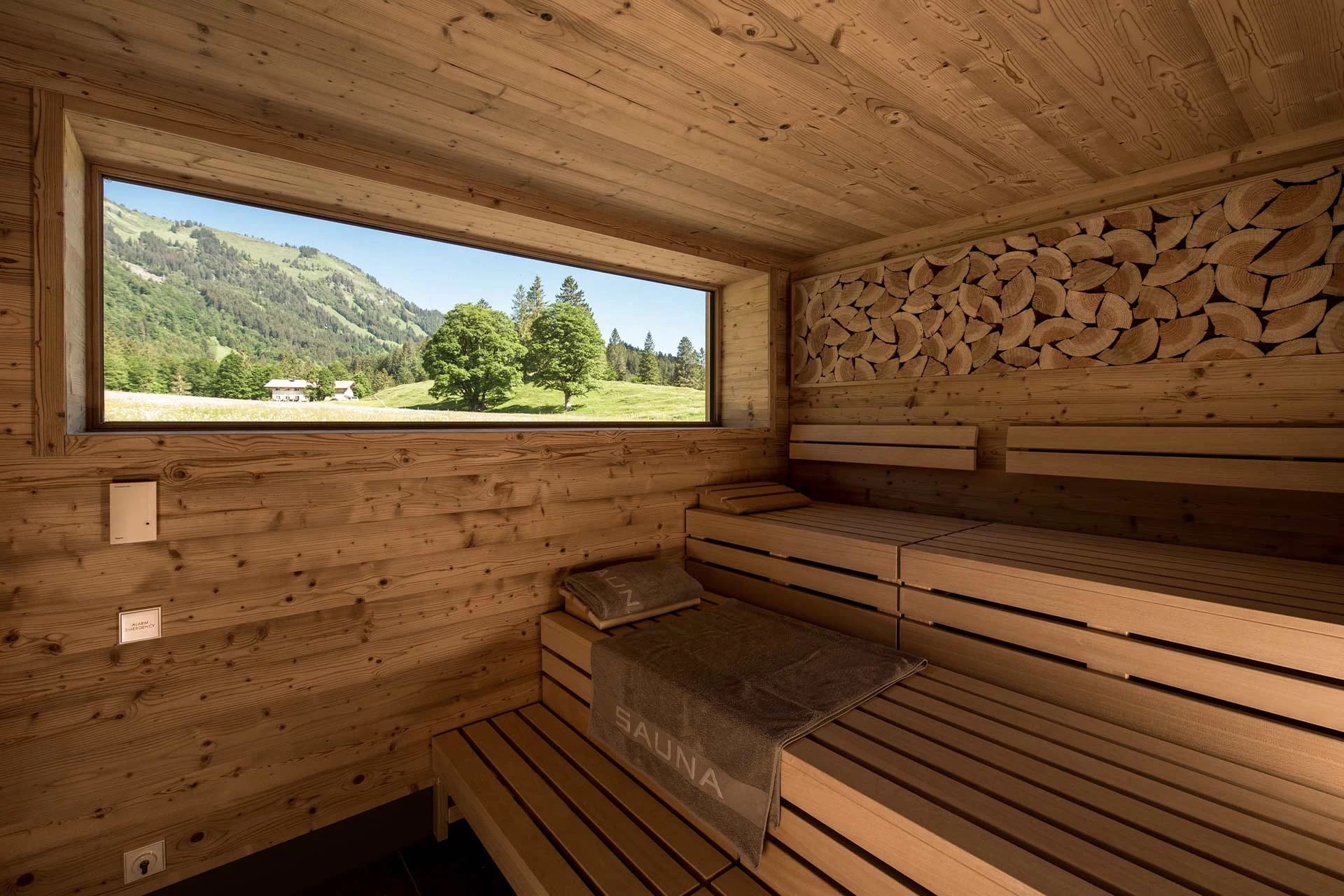 Sauna aus hellem Holz mit langem Panoramafenster und Blick auf Wiese und Berge