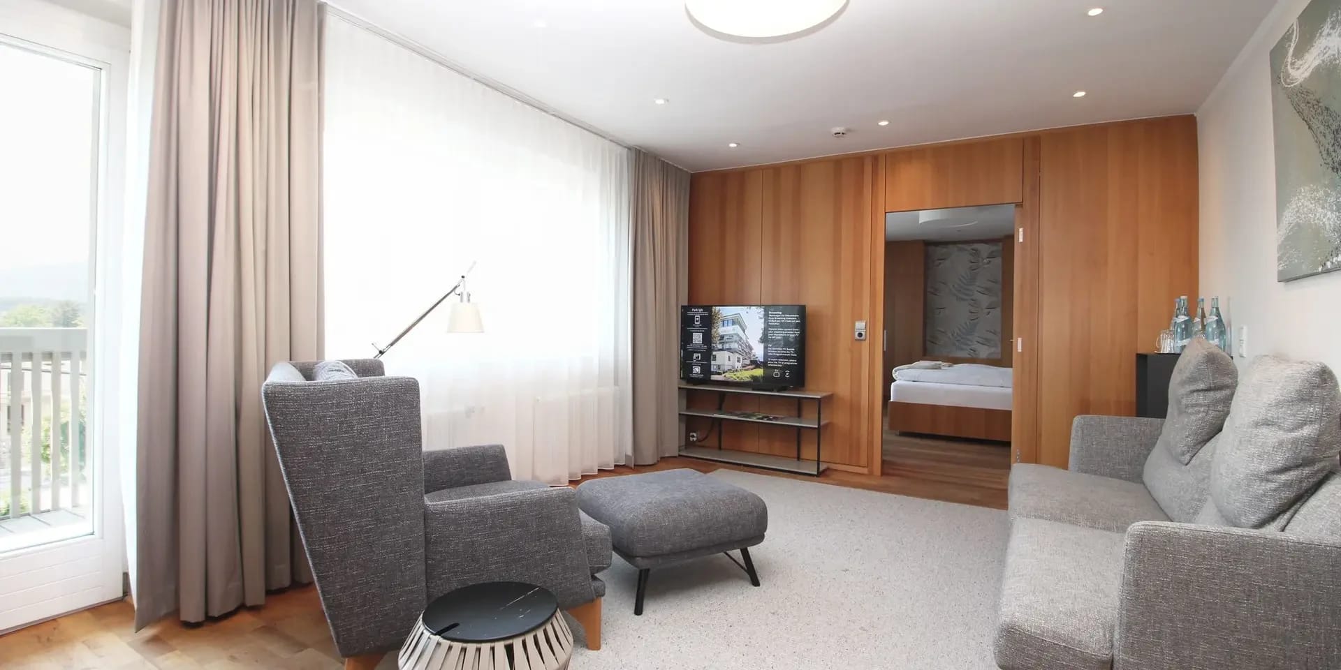 Moderner Wohnbereich mit grauen Sesseln, Hocker, Holzwand und offenem Durchgang zum Schlafzimmer