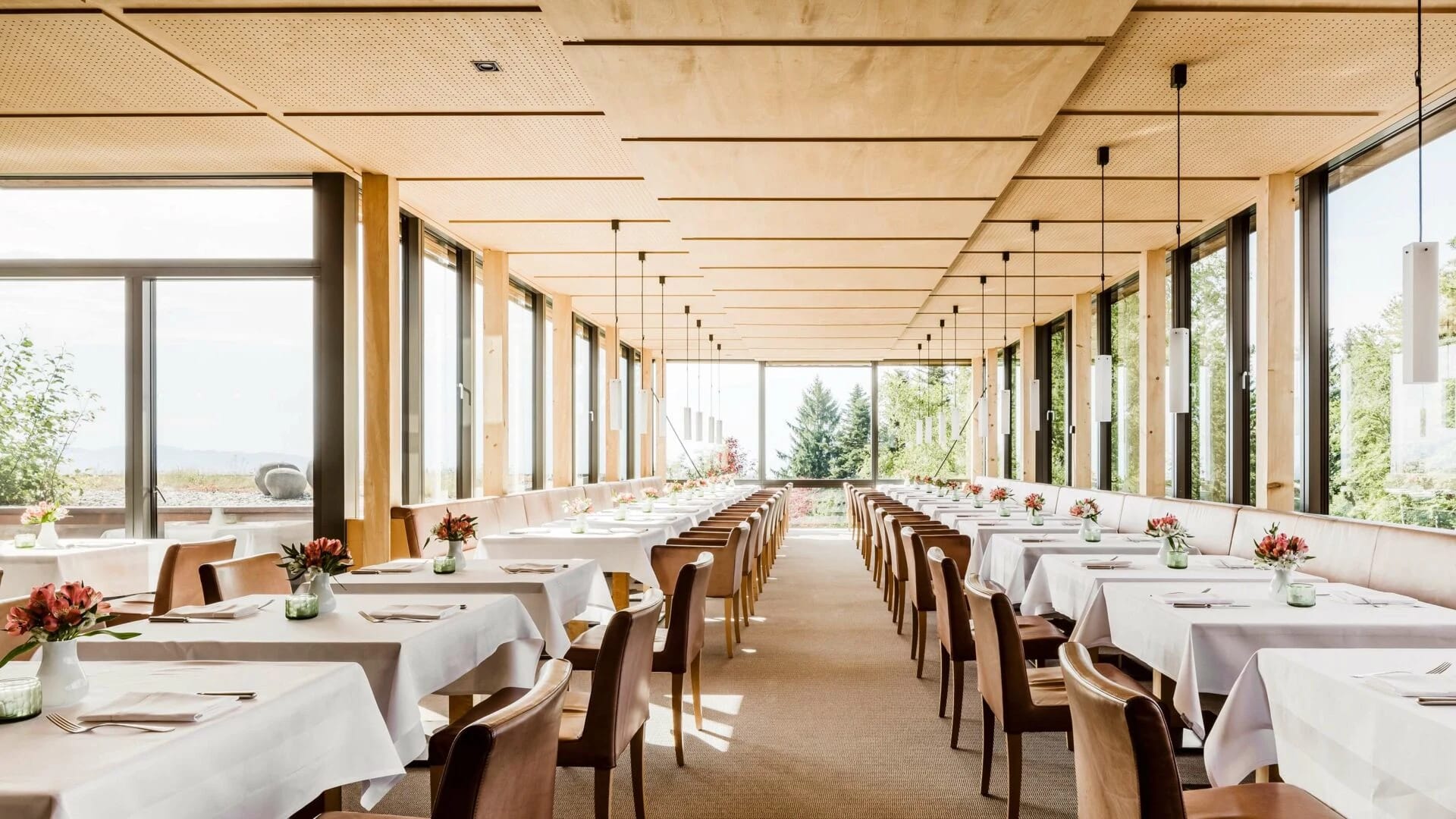Speisesaal mit langen, weiß eingedeckten Tischen, Holzdecke und Fensterfront