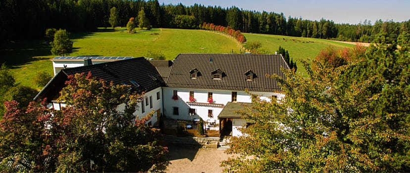 Außenansicht des weißen Landhotels mit Satteldach vor grünen Hügeln und Wald