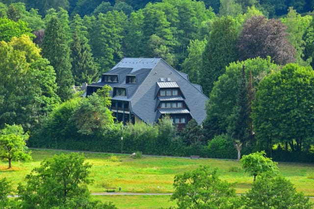 Das Hotel mit dunklem Mansarddach zwischen hohen Bäumen, davor eine grüne Wiese
