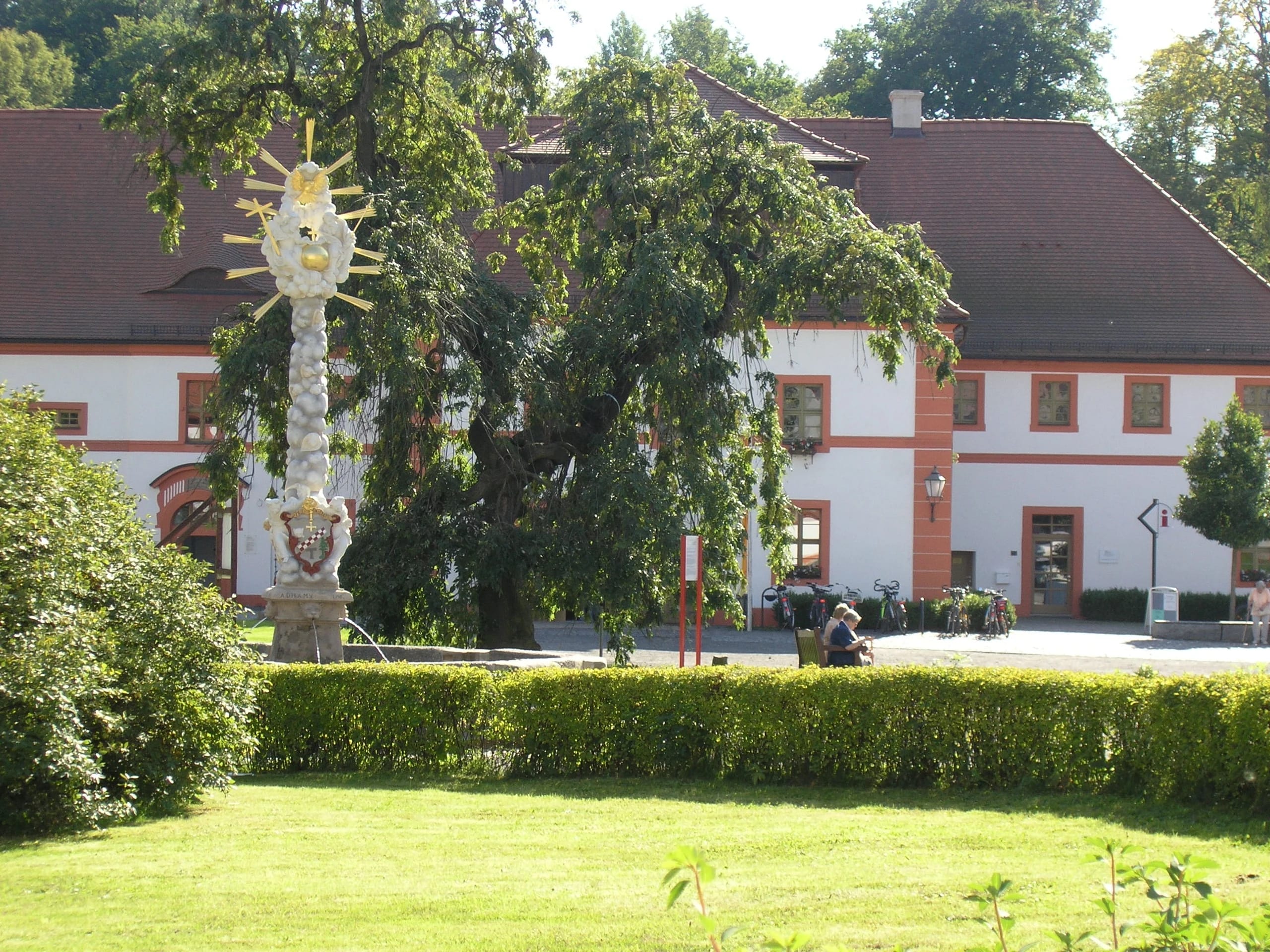 Klosterhof mit vergoldeter Dreifaltigkeitssäule, altem Baum und weiß-rotem Wirtschaftsgebäude
