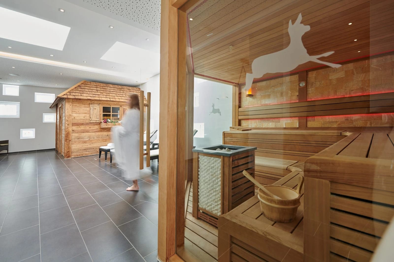 Sauna mit Holzbänken und Saunaofen, dahinter der Ruhebereich hinter Glas
