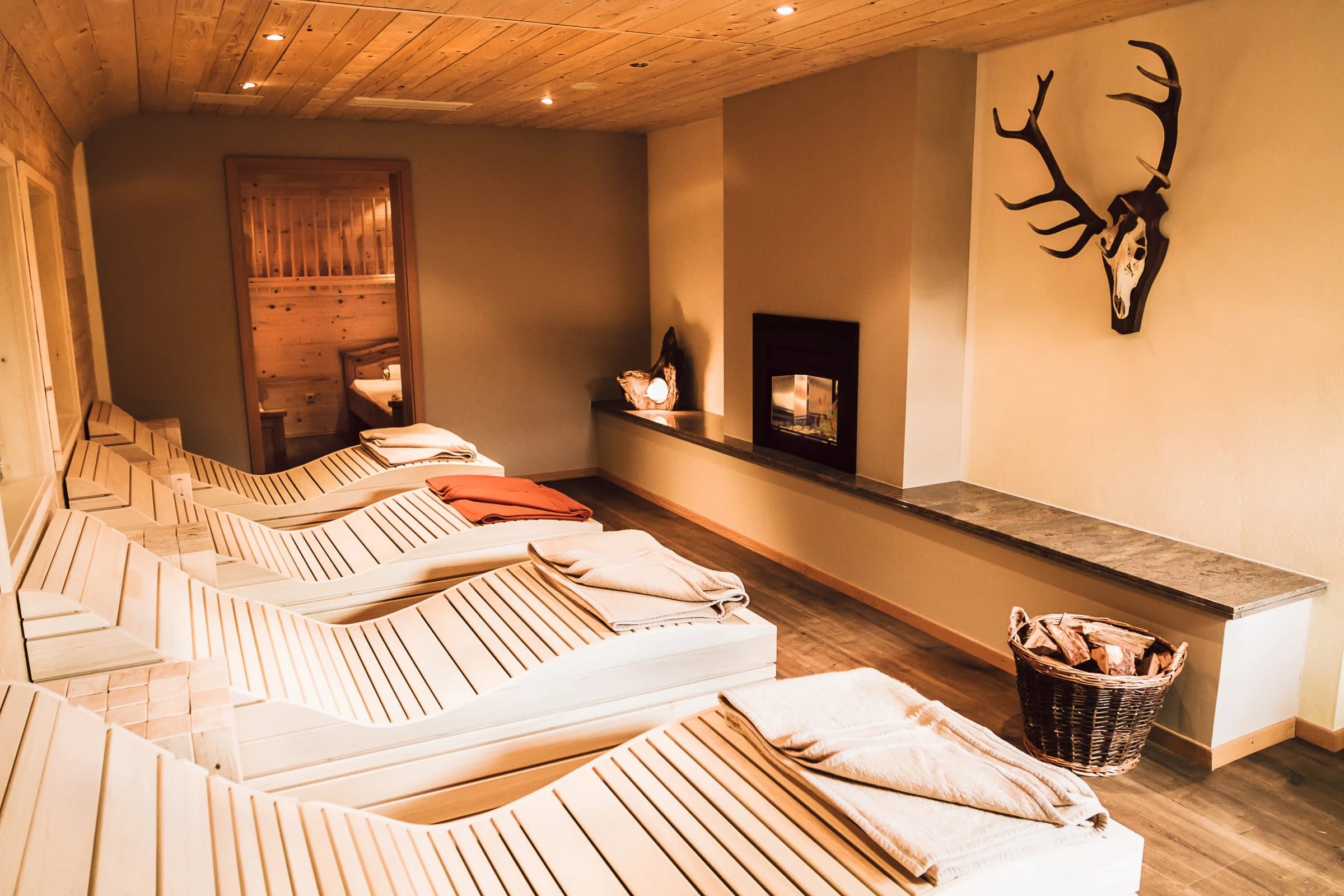 Relaxraum mit Holzliegen, Kaminfeuer und Geweih an der Wand