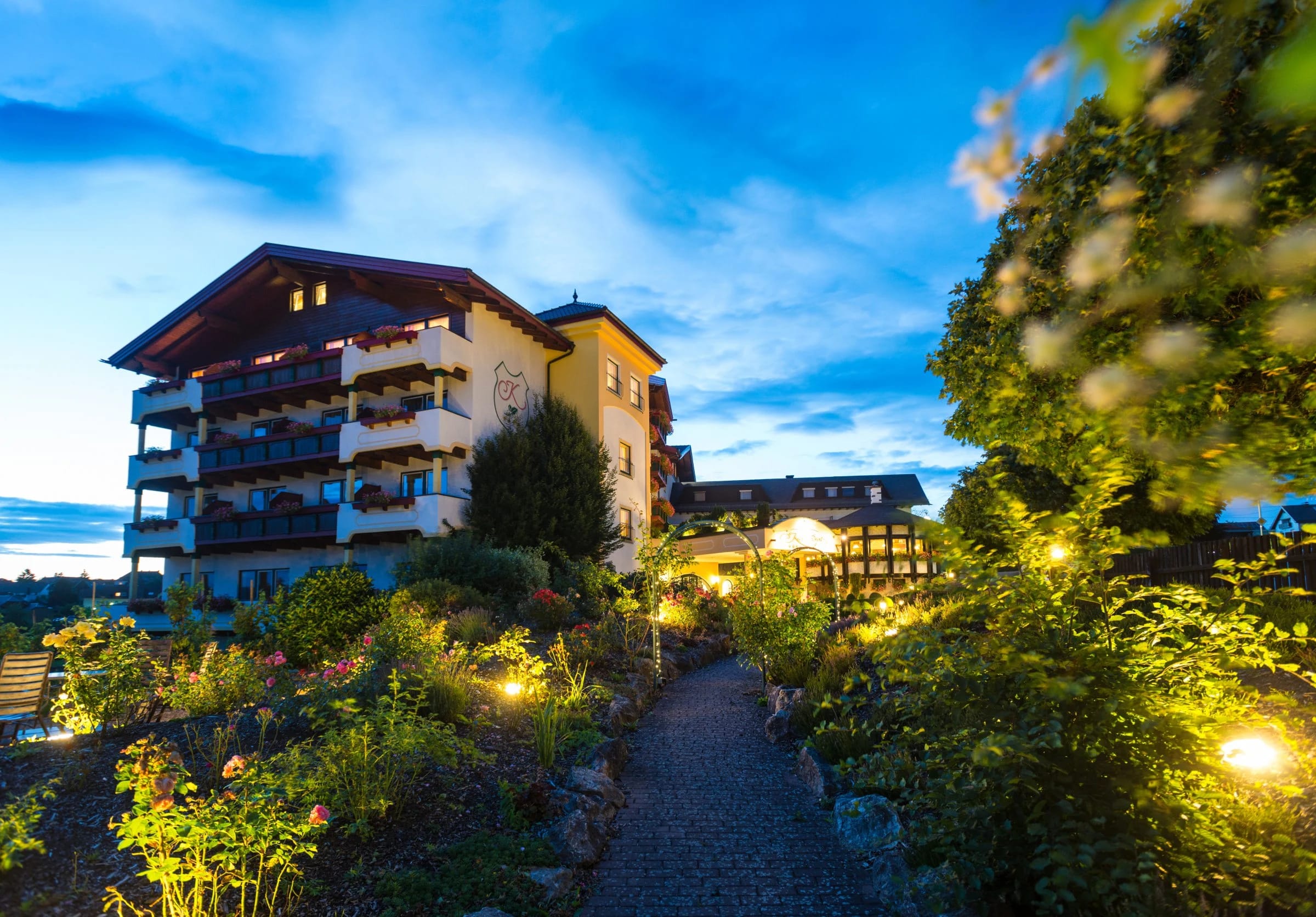 Hotel in der Abenddämmerung mit beleuchtetem Gartenweg und blühenden Beeten
