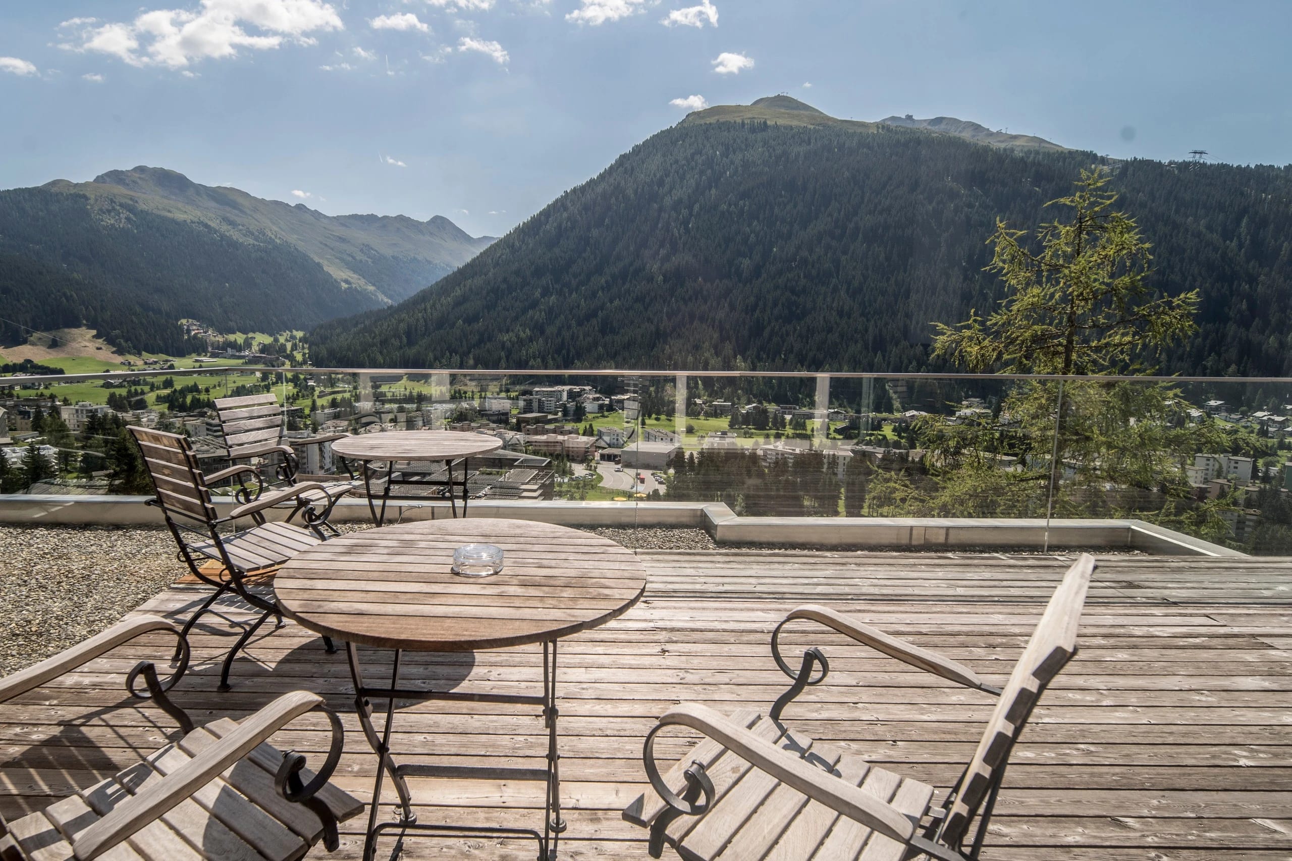 Holzterrasse mit runden Tischen und Gartenstühlen, Blick über das Tal auf bewaldete Berge