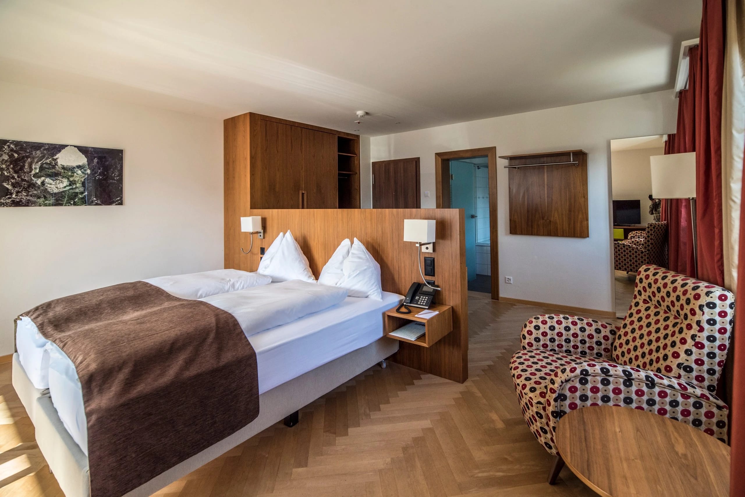 Doppelzimmer mit Holzkopfteil, brauner Tagesdecke, gemustertem Sessel und Parkettboden