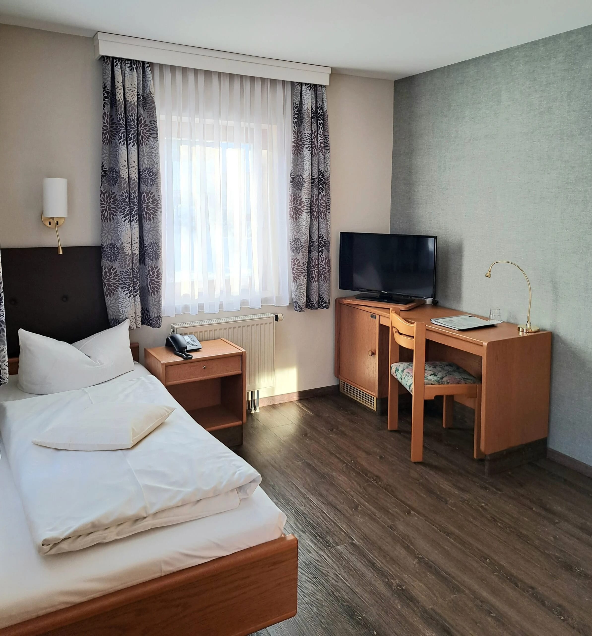 Hotelzimmer mit Bett, Schreibtisch, Fernseher und Fenster mit gemusterten Vorhängen