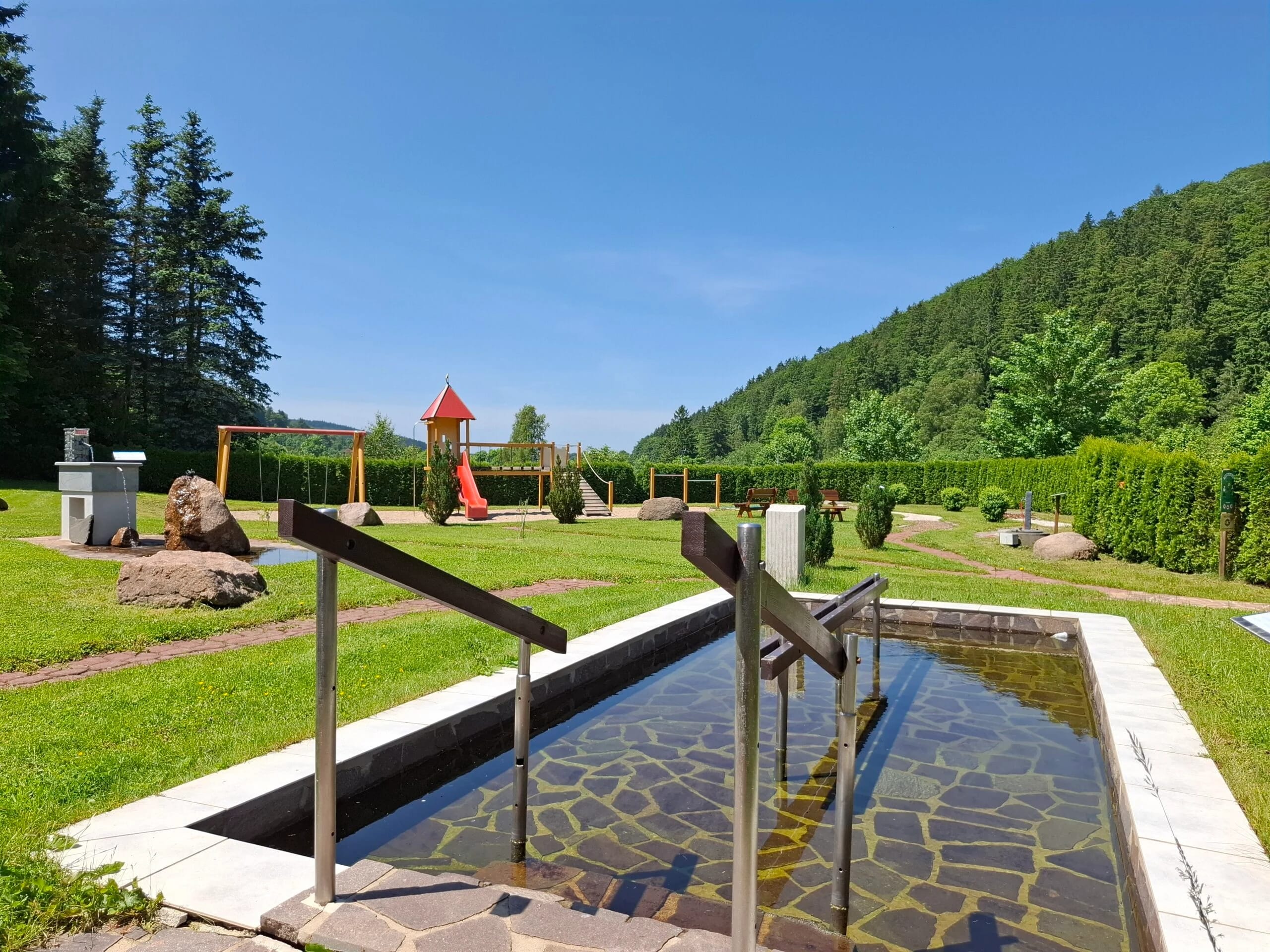 Wassertretbecken und Spielplatz auf der Wiese vor bewaldetem Hang