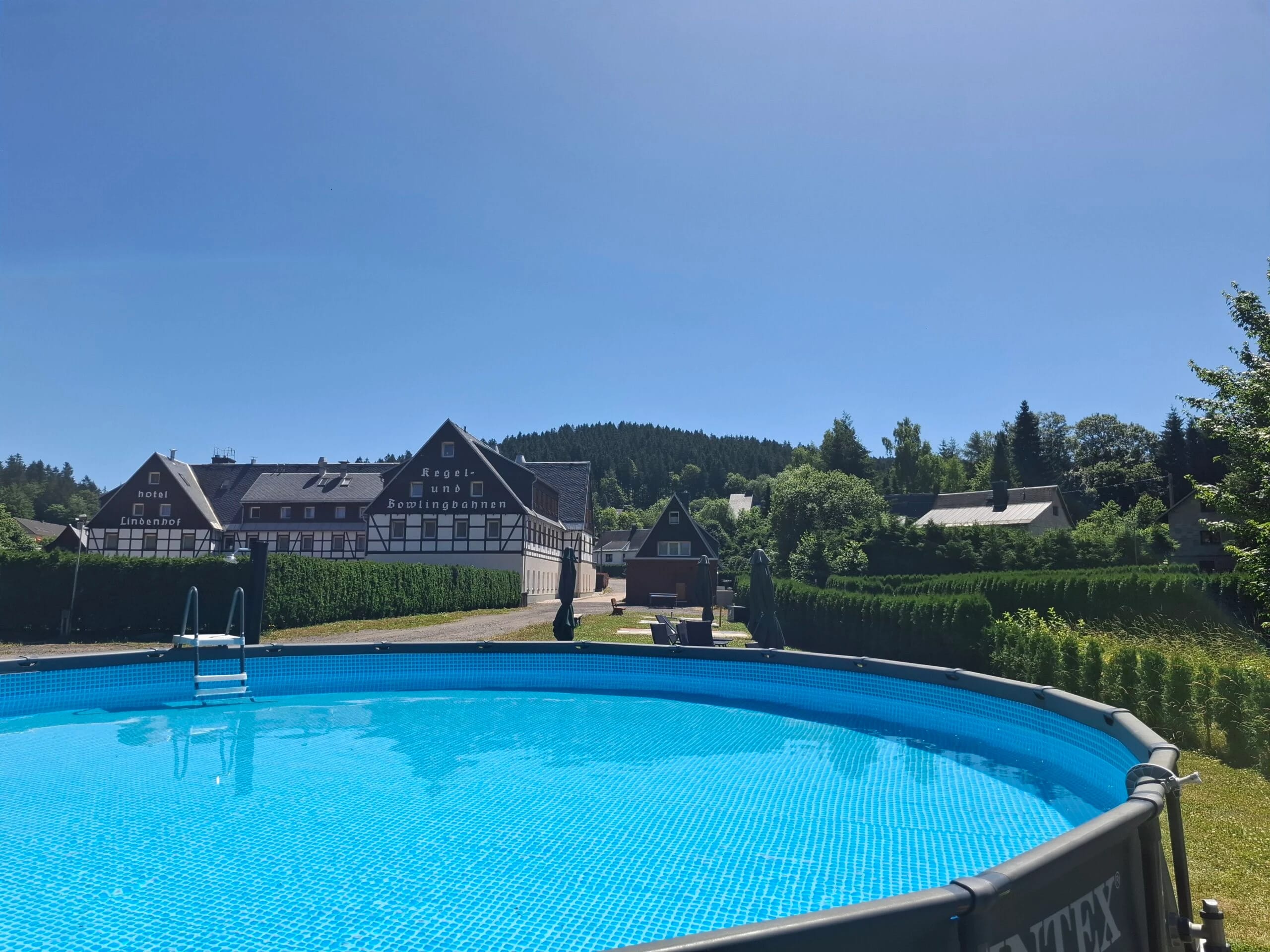 Runder Aufstellpool im Garten, dahinter das Fachwerkhaus des Hotels und bewaldete Hügel