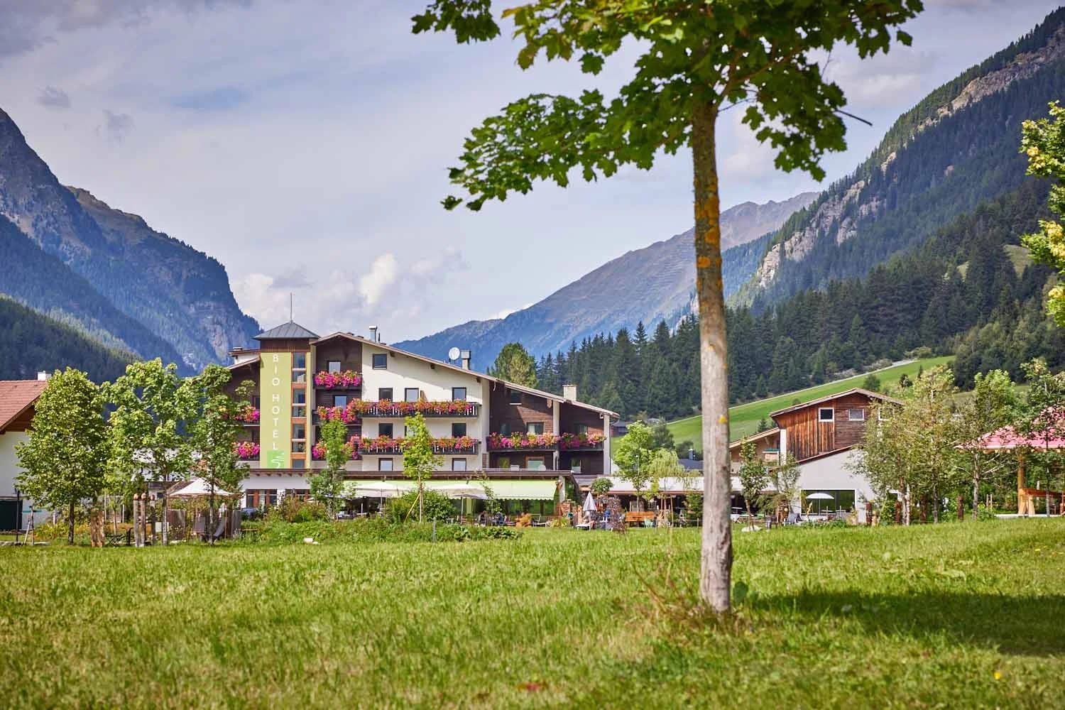 Hotelgebäude mit Blumenbalkonen, davor eine grüne Wiese und Berge im Hintergrund