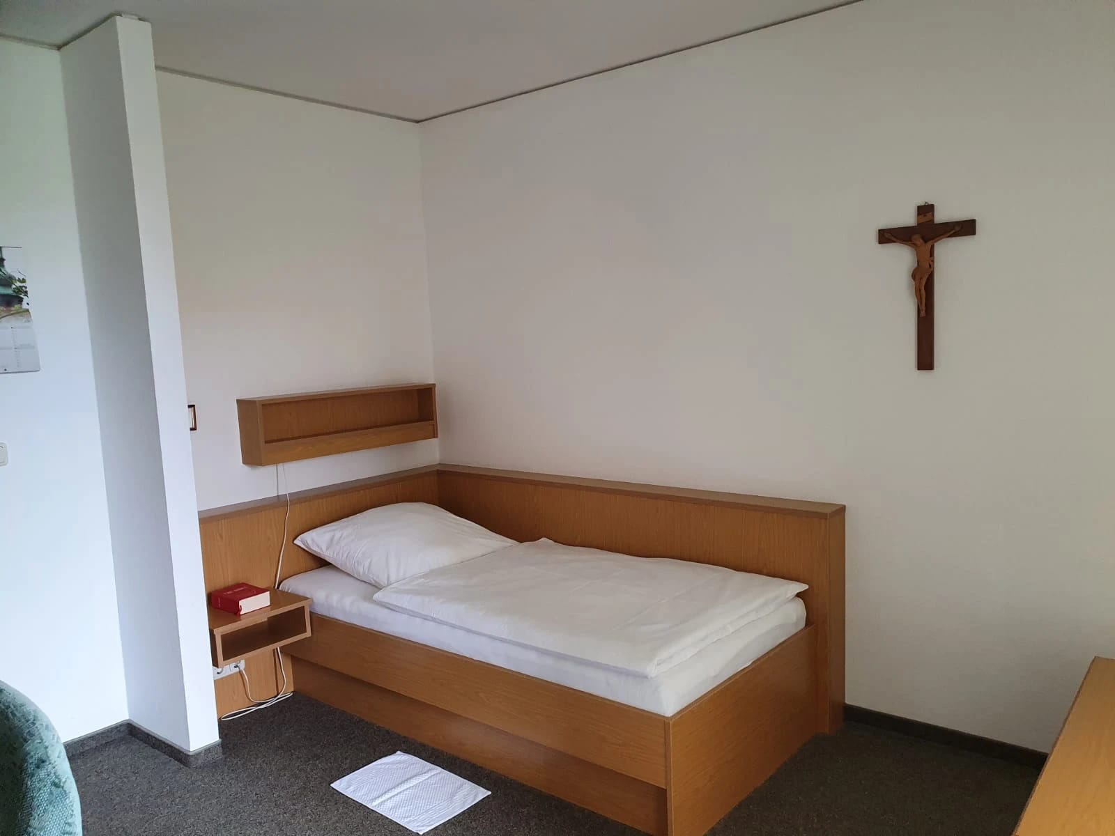 Einzelzimmer mit Holzbett, weißer Bettwäsche, Nachttisch und Kruzifix an der Wand