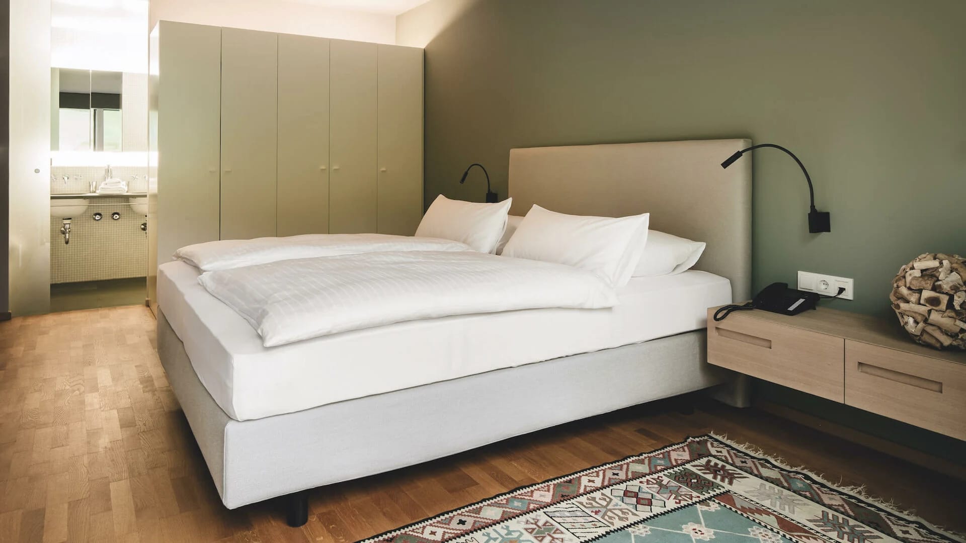 Doppelzimmer mit grüner Wand, Holzboden, Nachttisch und Blick ins Bad