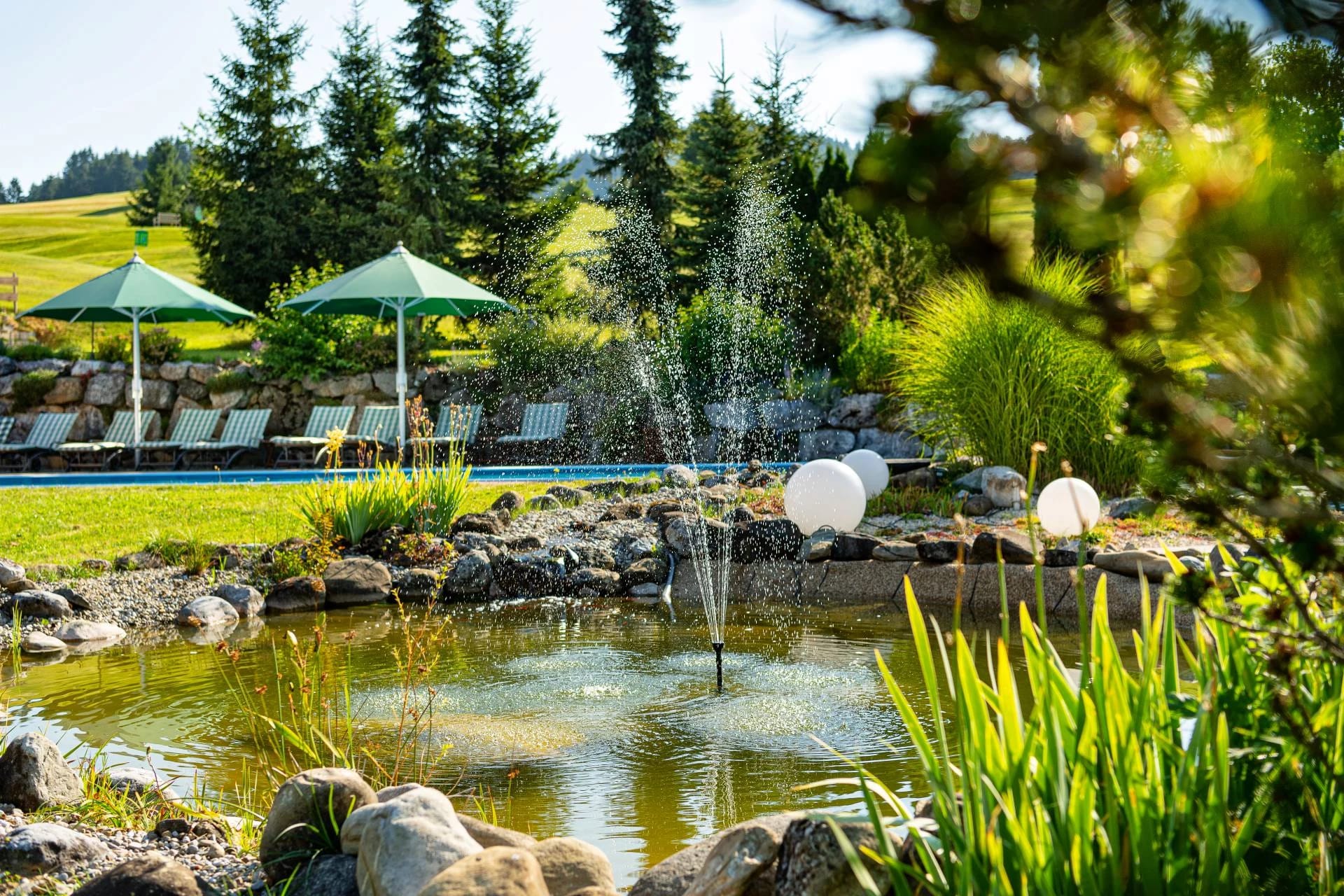 Gartenteich mit Springbrunnen, dahinter Sonnenliegen am Pool