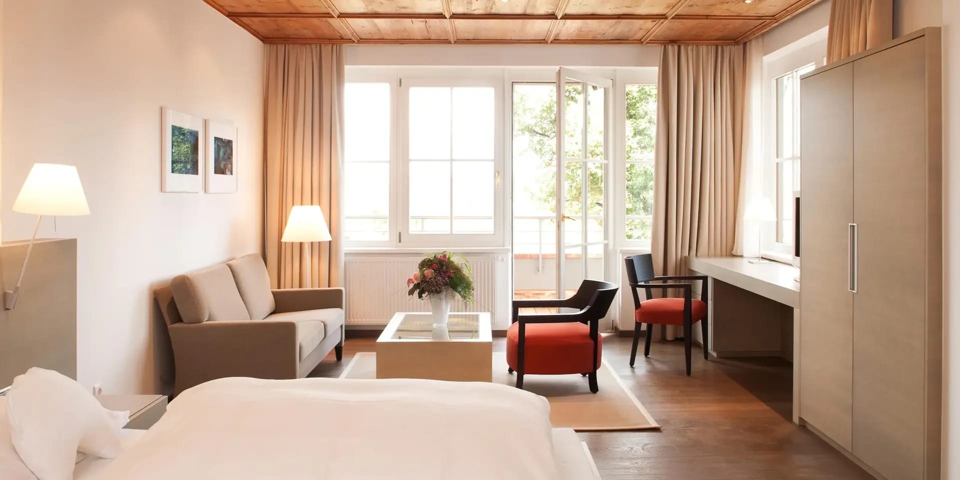 Zimmer mit Holzdecke, Doppelbett, Sofa, Sesseln und Schreibtisch vor bodentiefen Fenstern
