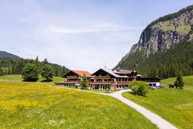 Hotelgebäude in einer blühenden Bergwiese, dahinter Wald und eine steile Felswand