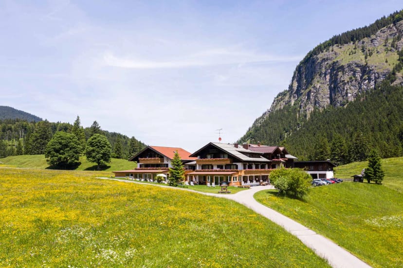 Hotelgebäude in einer blühenden Bergwiese, dahinter Wald und eine steile Felswand
