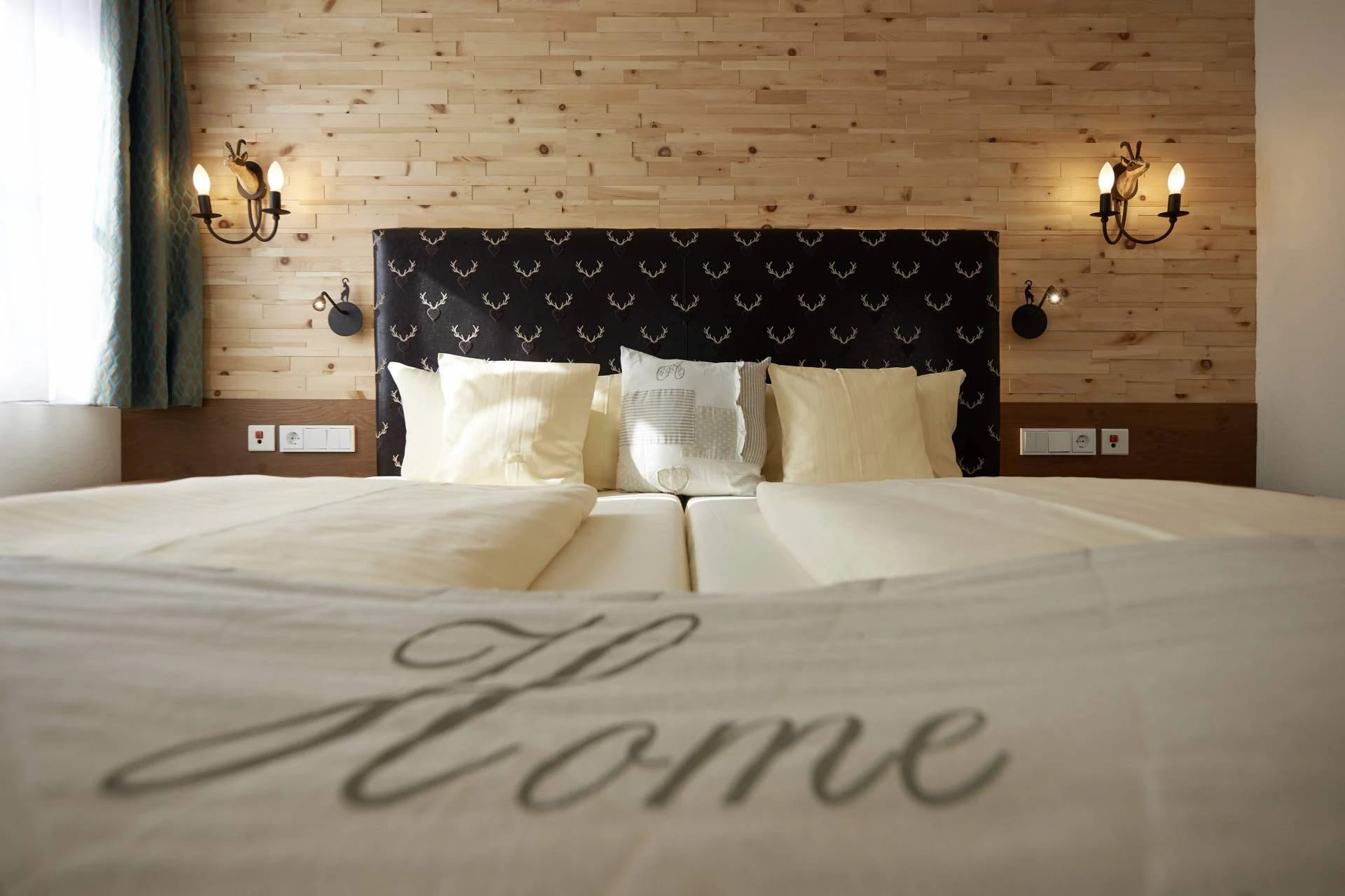 Frisch bezogenes Doppelbett mit dunklem Kopfteil, heller Holzwand und zwei Wandleuchten
