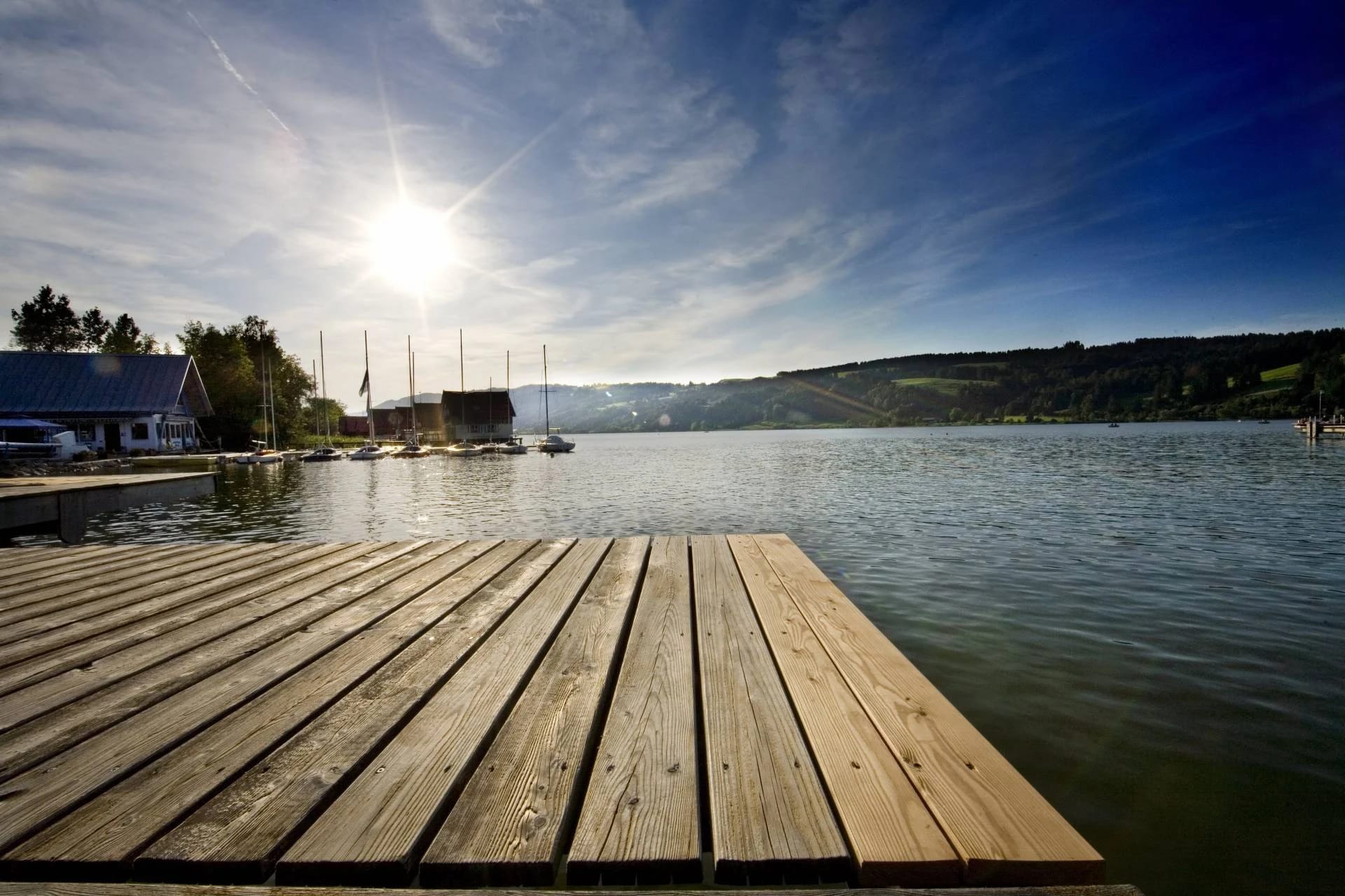 Holzsteg an einem See mit Segelbooten, Sonne und bewaldeten Hügeln im Hintergrund