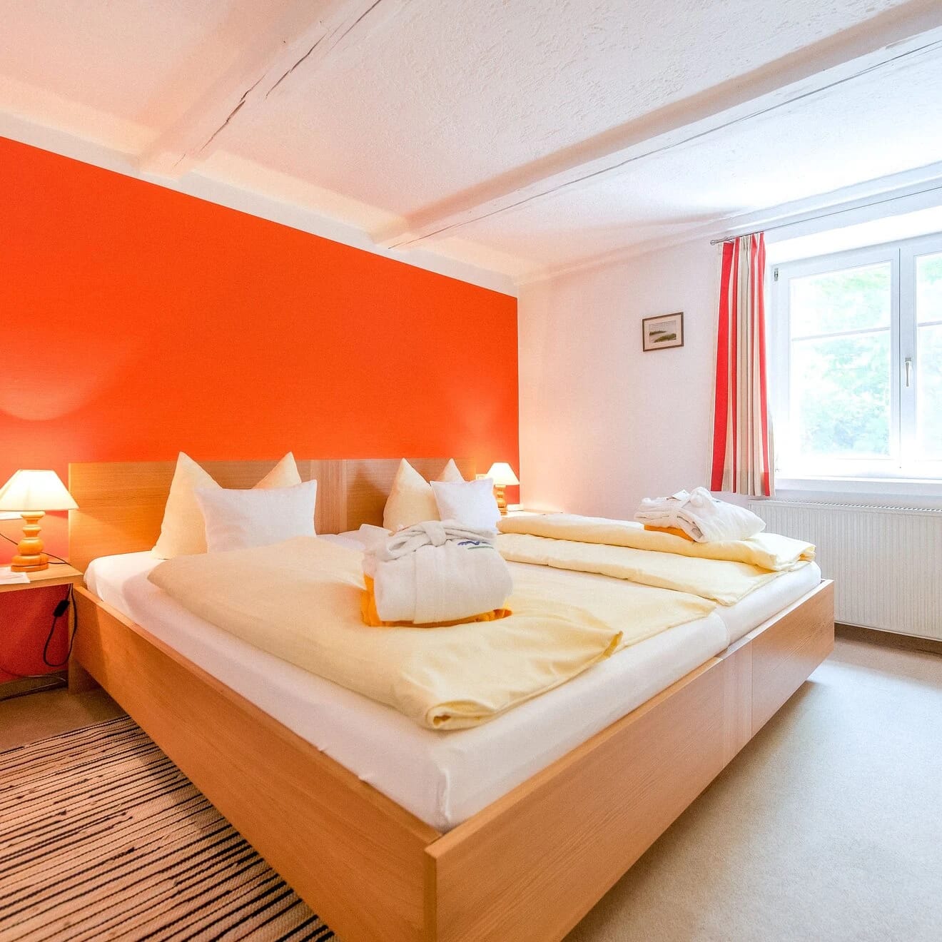Doppelzimmer mit Holzbett vor orangefarbener Wand und Fenster mit gestreiften Vorhängen