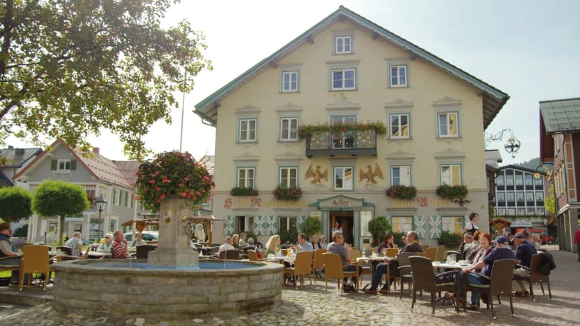 Hellgelbe Hausfassade mit Adler-Malerei und Blumenkästen, davor ein Steinbrunnen und Gäste an Tischen im Freien