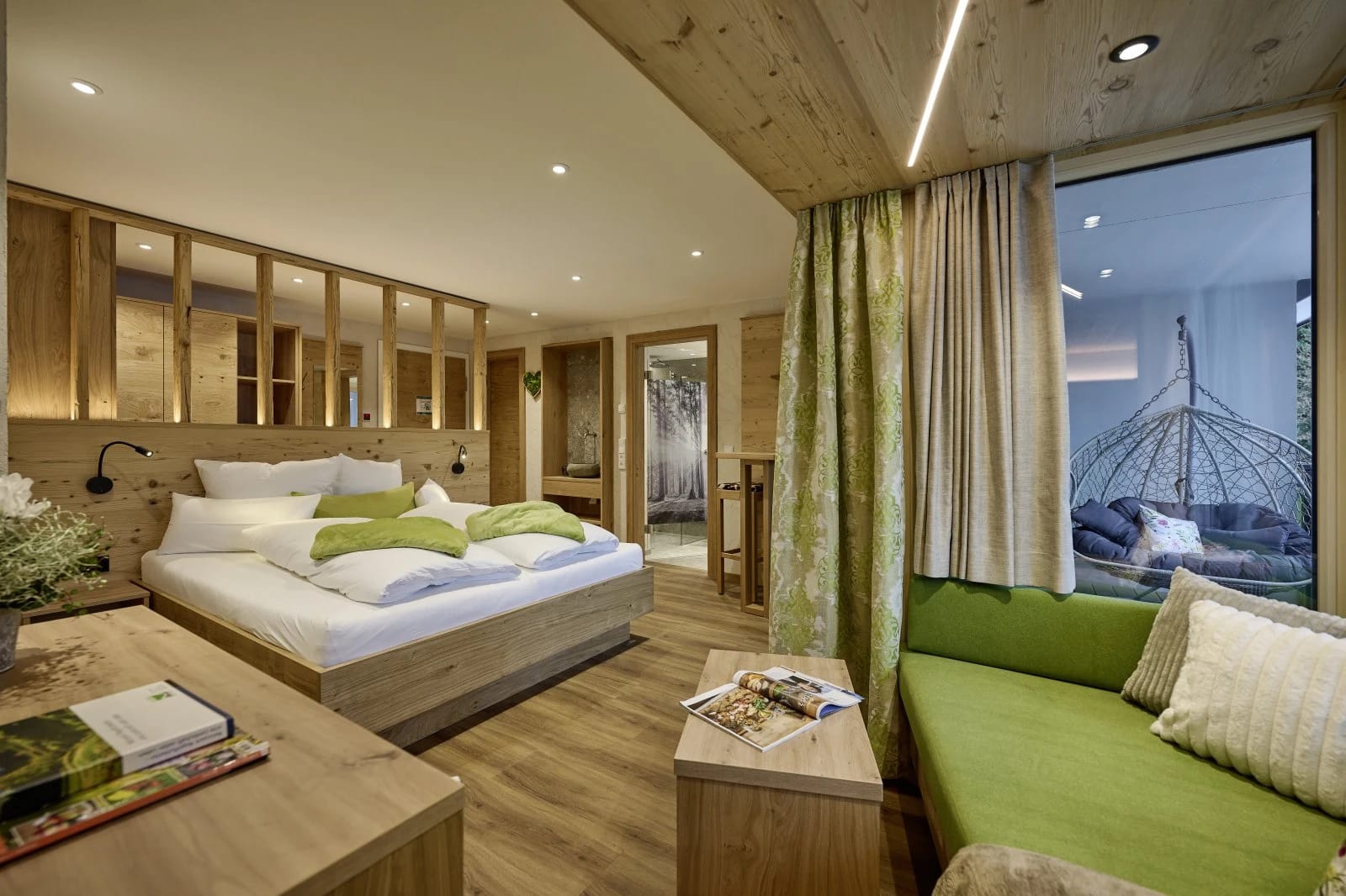 Zimmer mit Doppelbett, grünem Sofa und Hängesessel hinter einer Glaswand
