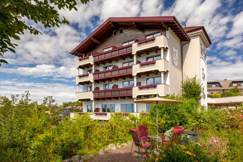 Hotelgebäude mit Holzbalkonen, davor Gartenterrasse mit Stühlen und Sonnenschirm