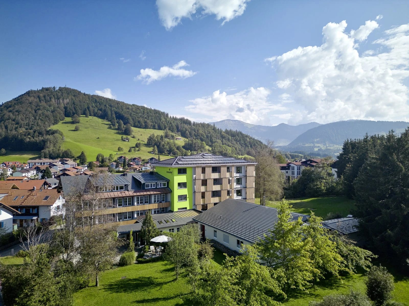 Luftaufnahme des Hotels mit begrünter Fassade, Ortschaft und Bergpanorama im Hintergrund