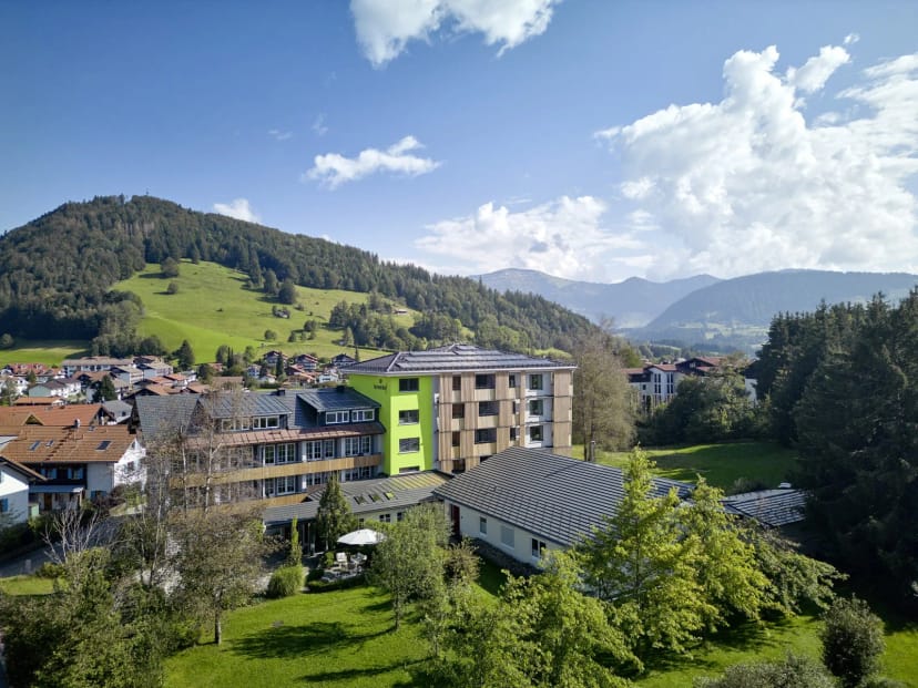 Luftaufnahme des Hotels mit begrünter Fassade, Ortschaft und Bergpanorama im Hintergrund