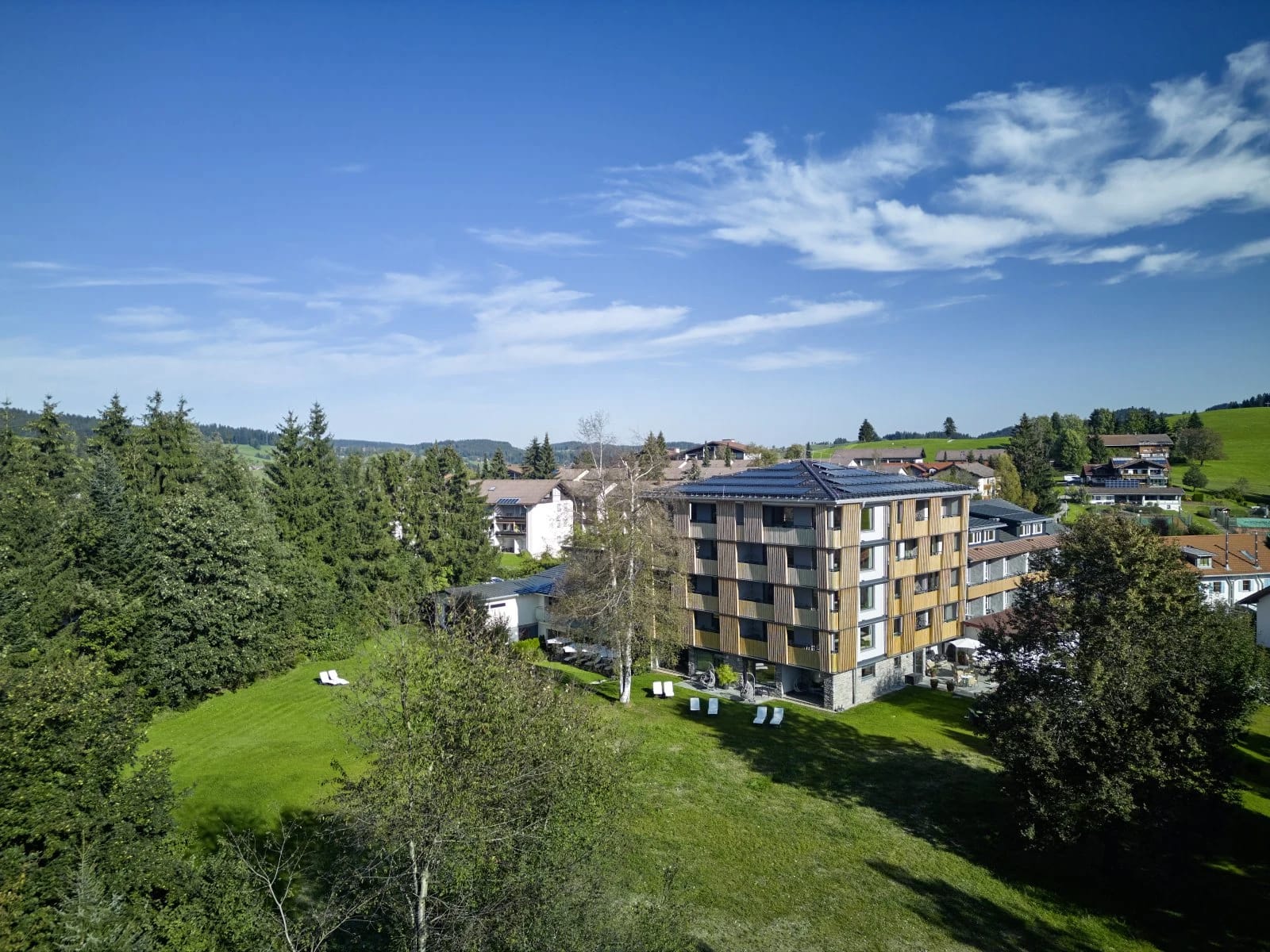 Das Hotelgebäude von der Gartenseite mit Liegewiese, Bäumen und blauem Himmel