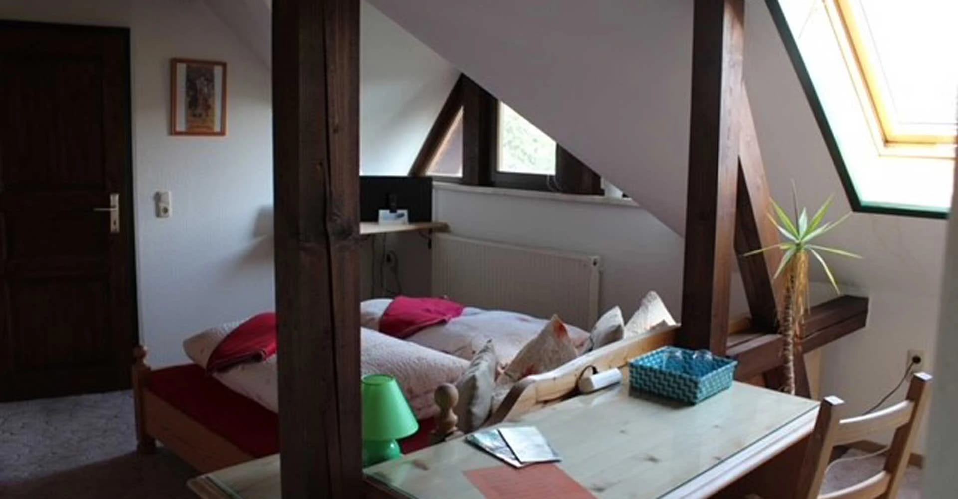 Dachzimmer mit sichtbaren Holzbalken, Bett mit bunten Kissen und Sitzecke am Dachfenster