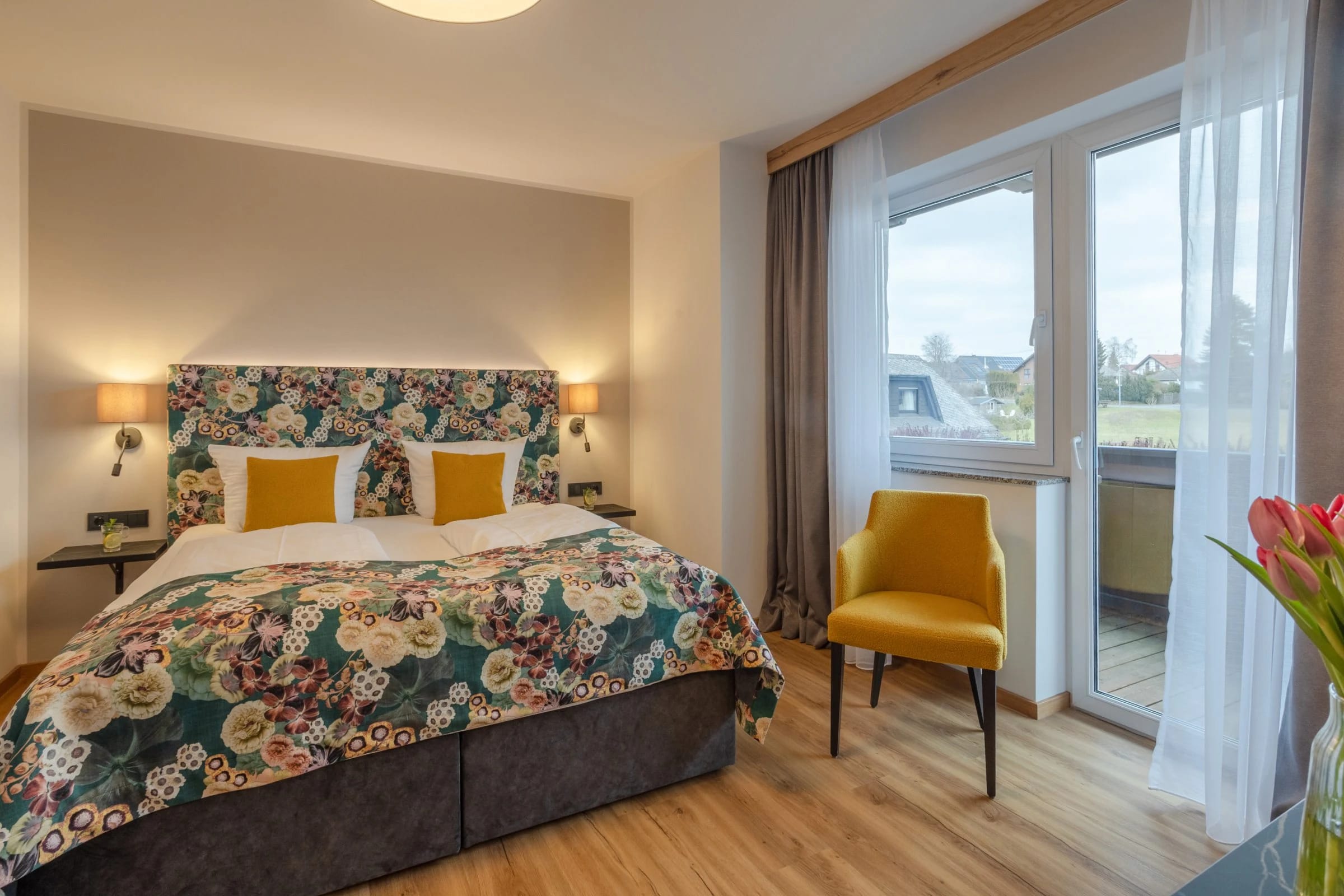 Zimmer mit blumigem Bettüberwurf, gelbem Sessel und Balkontür