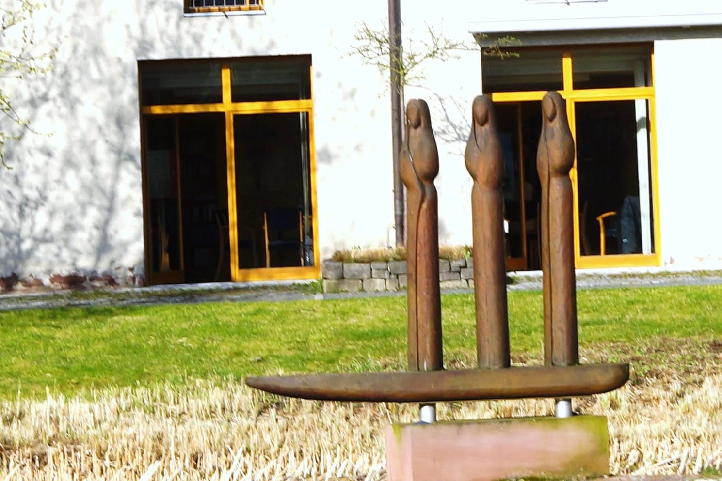 Rostbraune Holzskulptur mit drei Figuren auf der Wiese vor dem Gebäude mit gelb gerahmten Fenstern