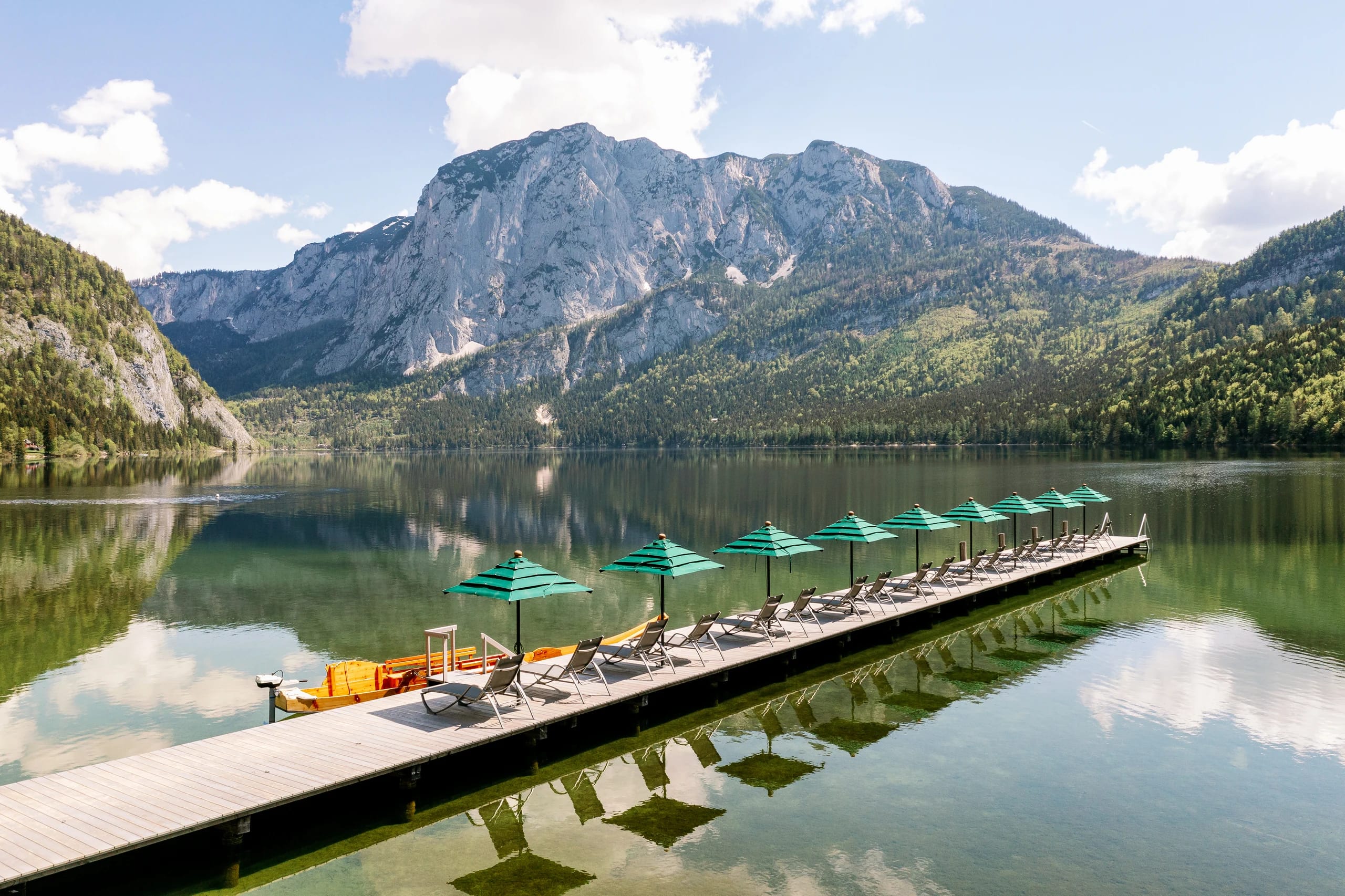 Steg mit grünen Sonnenschirmen und Liegen am See vor einem Bergmassiv