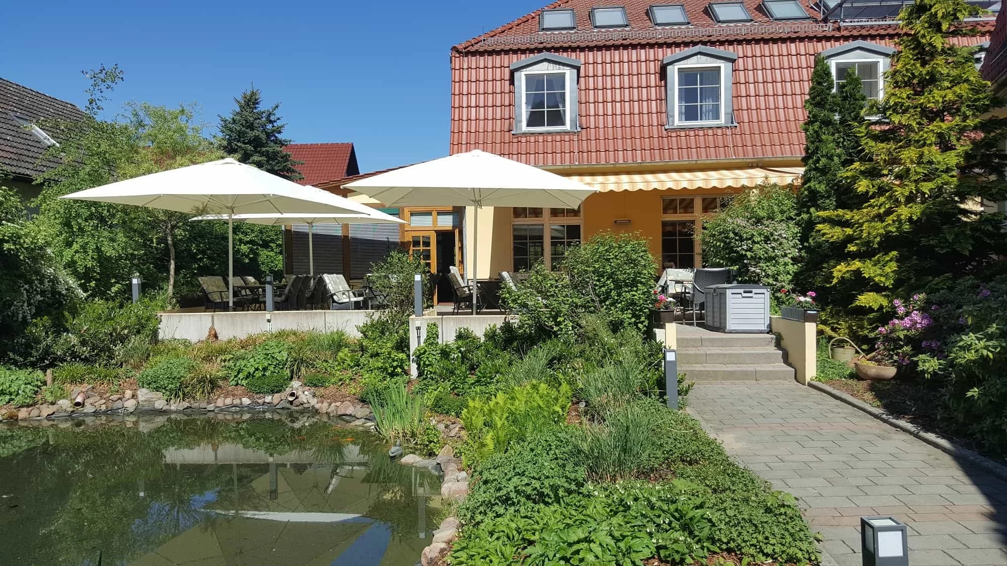 Sonnenterrasse mit Sonnenschirmen vor dem Haus, davor ein Gartenteich