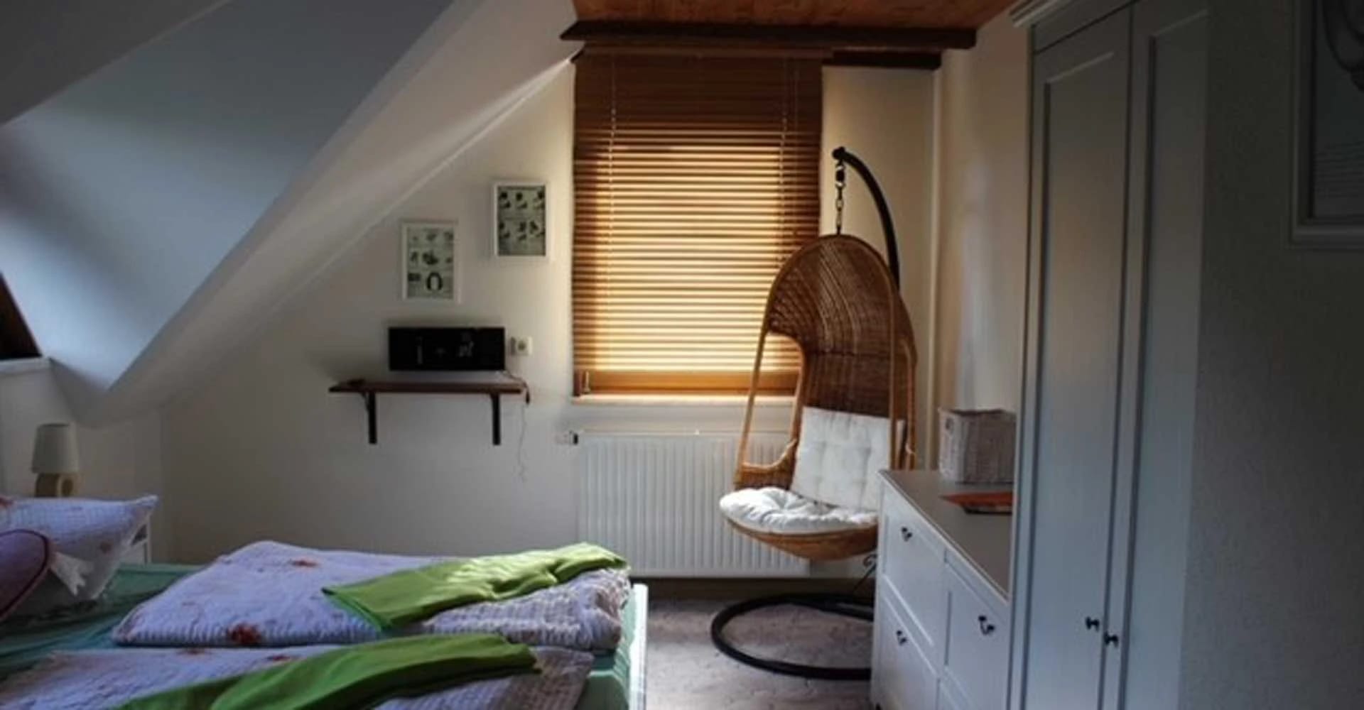 Dachzimmer mit Bett, Hängesessel aus Korbgeflecht und Fenster mit Holzjalousie