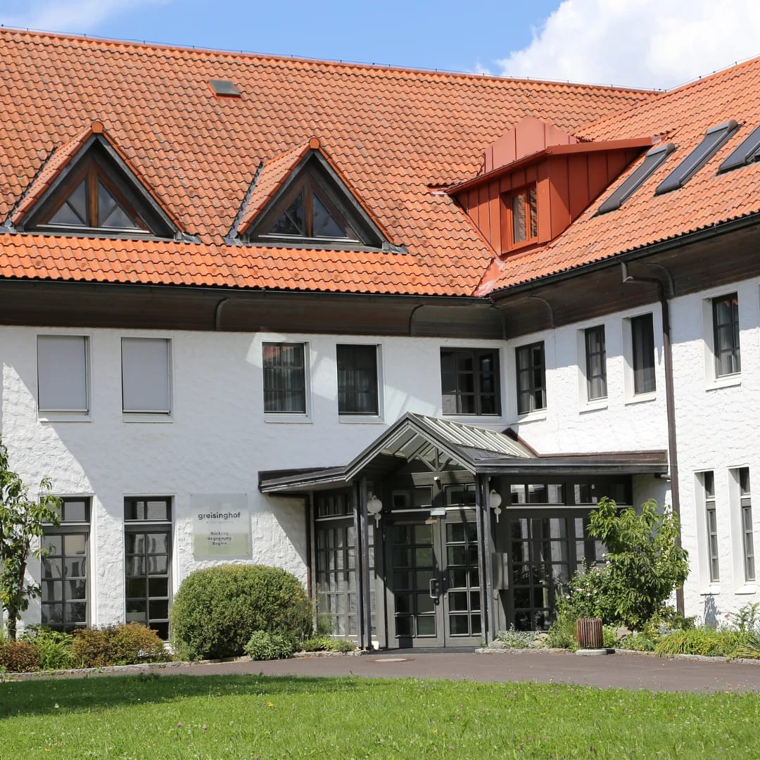 Weiß verputztes Haupthaus mit rotem Ziegeldach, Dachgaupen und verglastem Eingangsvorbau, davor Rasenfläche