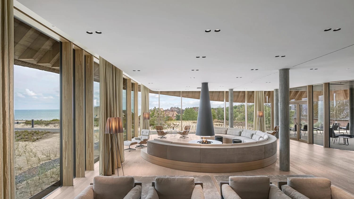 Lounge mit rundem Sitzbereich um einen offenen Kamin und bodentiefen Fenstern mit Blick über die Dünen