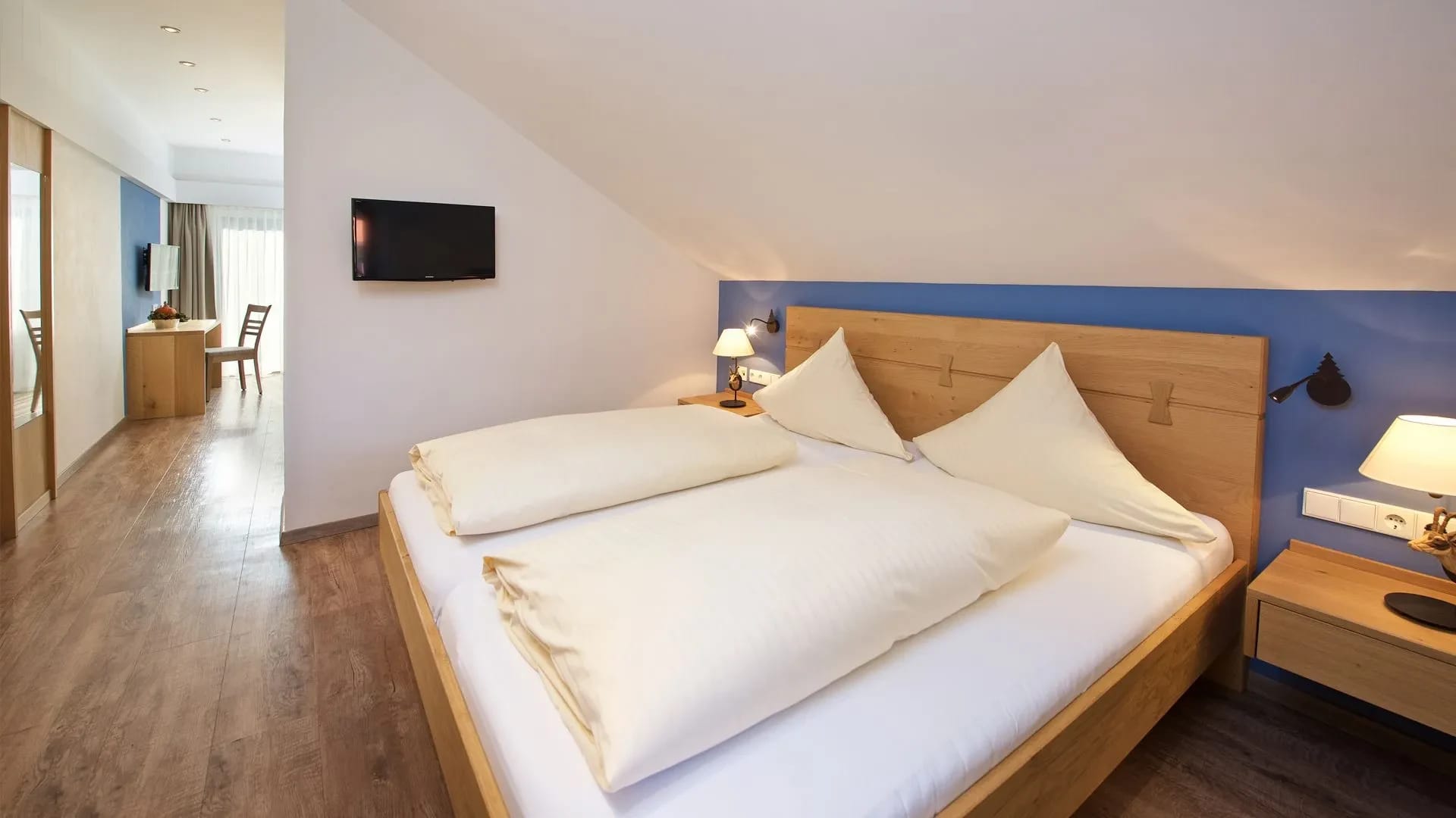 Doppelzimmer unter Dachschräge mit blauer Akzentwand, Holzbett, Wandfernseher und Sitzecke im Hintergrund