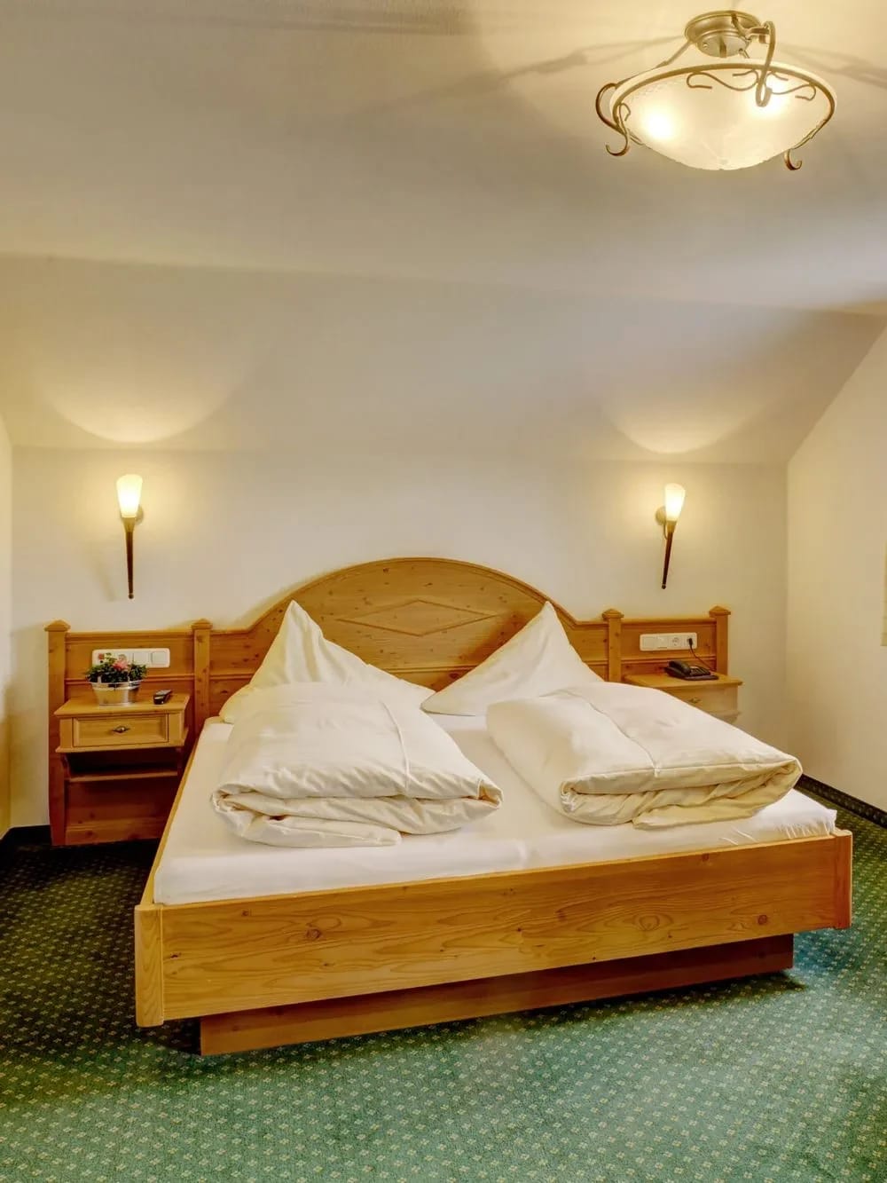 Doppelzimmer mit geschnitztem Holzbett, Nachttischen mit Wandleuchten und grünem Teppichboden