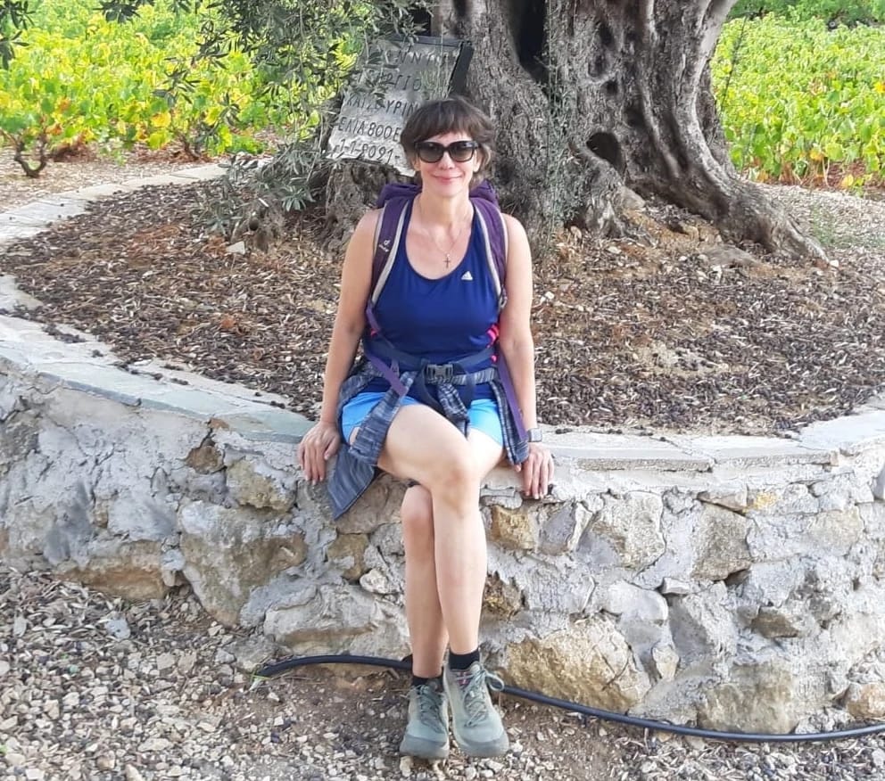 Dr. Nicola Stein sitzt in Wanderkleidung mit Sonnenbrille auf einer Steinmauer unter einem Olivenbaum