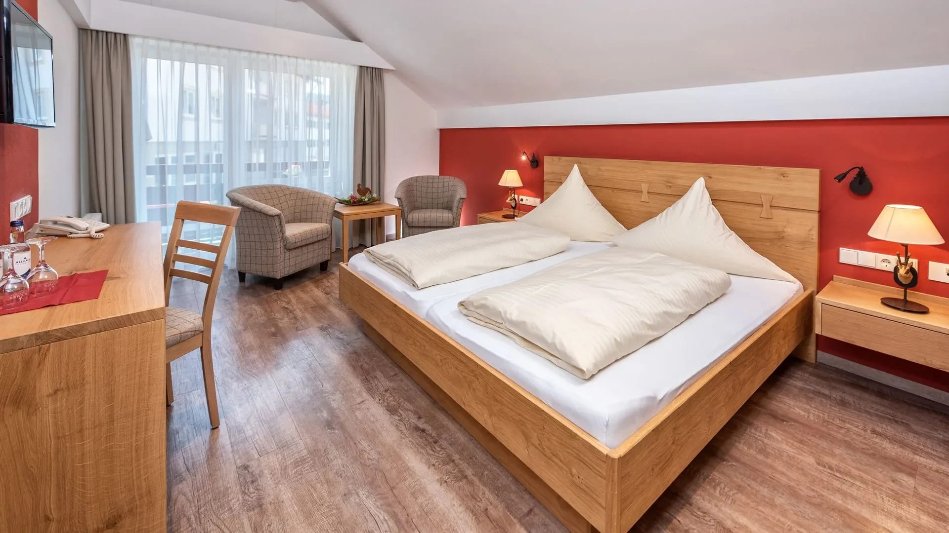 Doppelzimmer mit roter Akzentwand, Holzbett, Sesselgruppe am Fenster und Schreibtisch