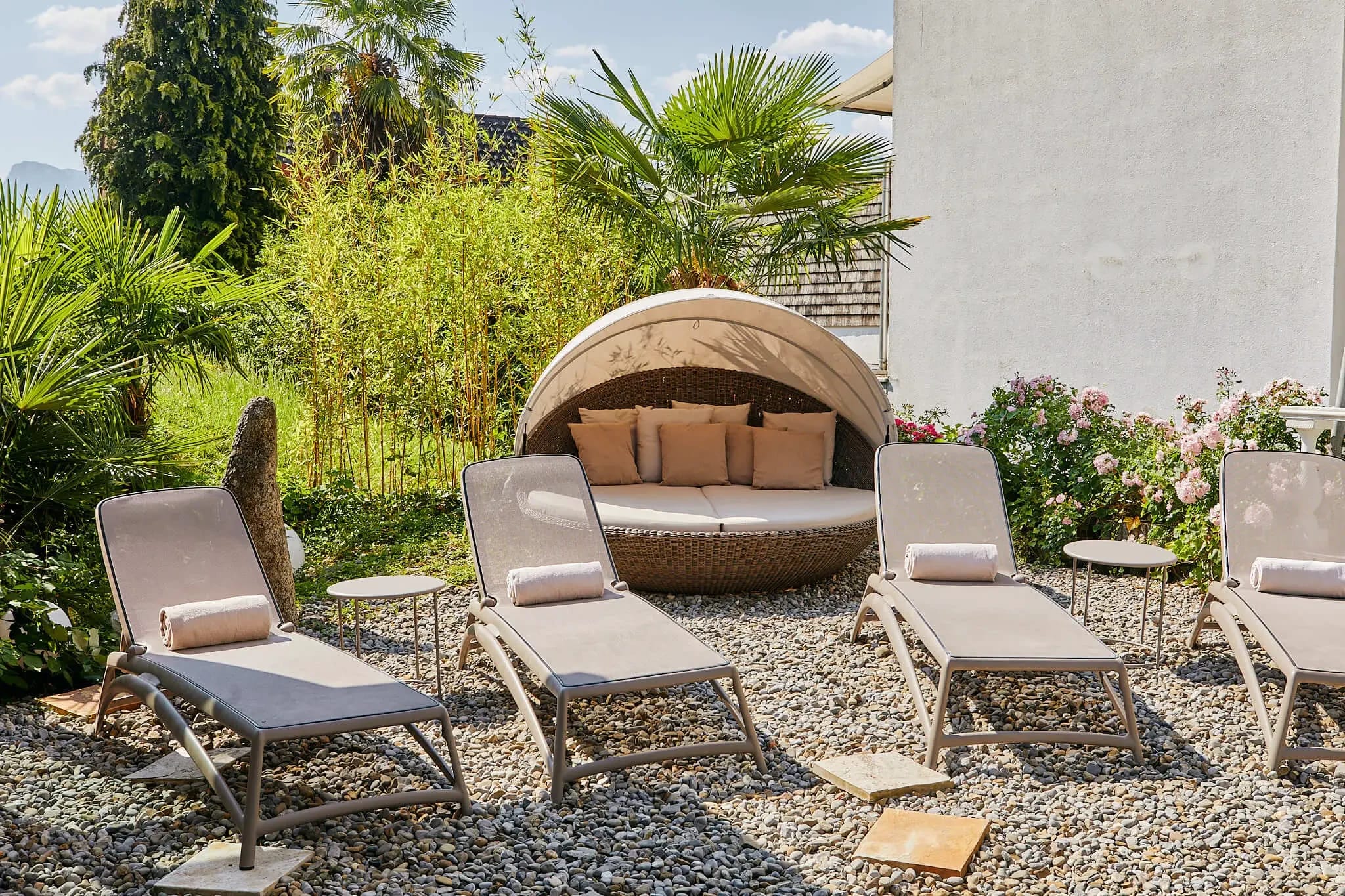 Liegestühle und eine runde Rattan-Muschelliege im Kiesgarten mit Palmen und blühenden Sträuchern