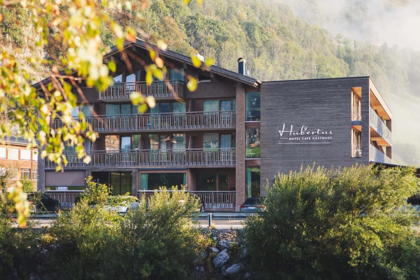 Außenansicht des Hotels mit Holzfassade und Balkonen vor bewaldetem Berghang, im Vordergrund herbstliches Laub
